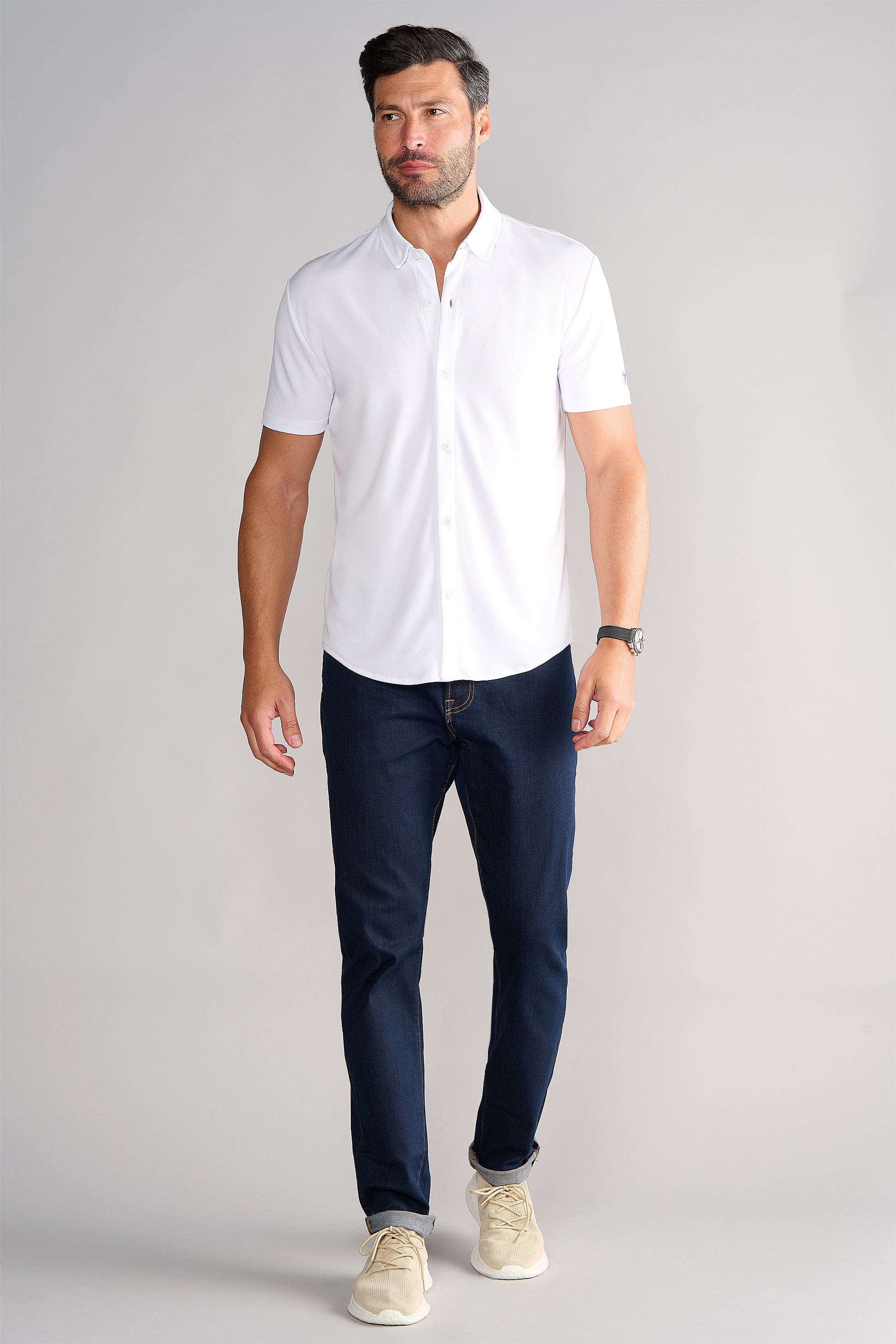 A man with short dark hair and a beard wears the Twillory Short Sleeve Button Down Polo Solid in white, dark blue jeans, beige sneakers, and a wristwatch. He stands against a light gray background, looking slightly left while walking.