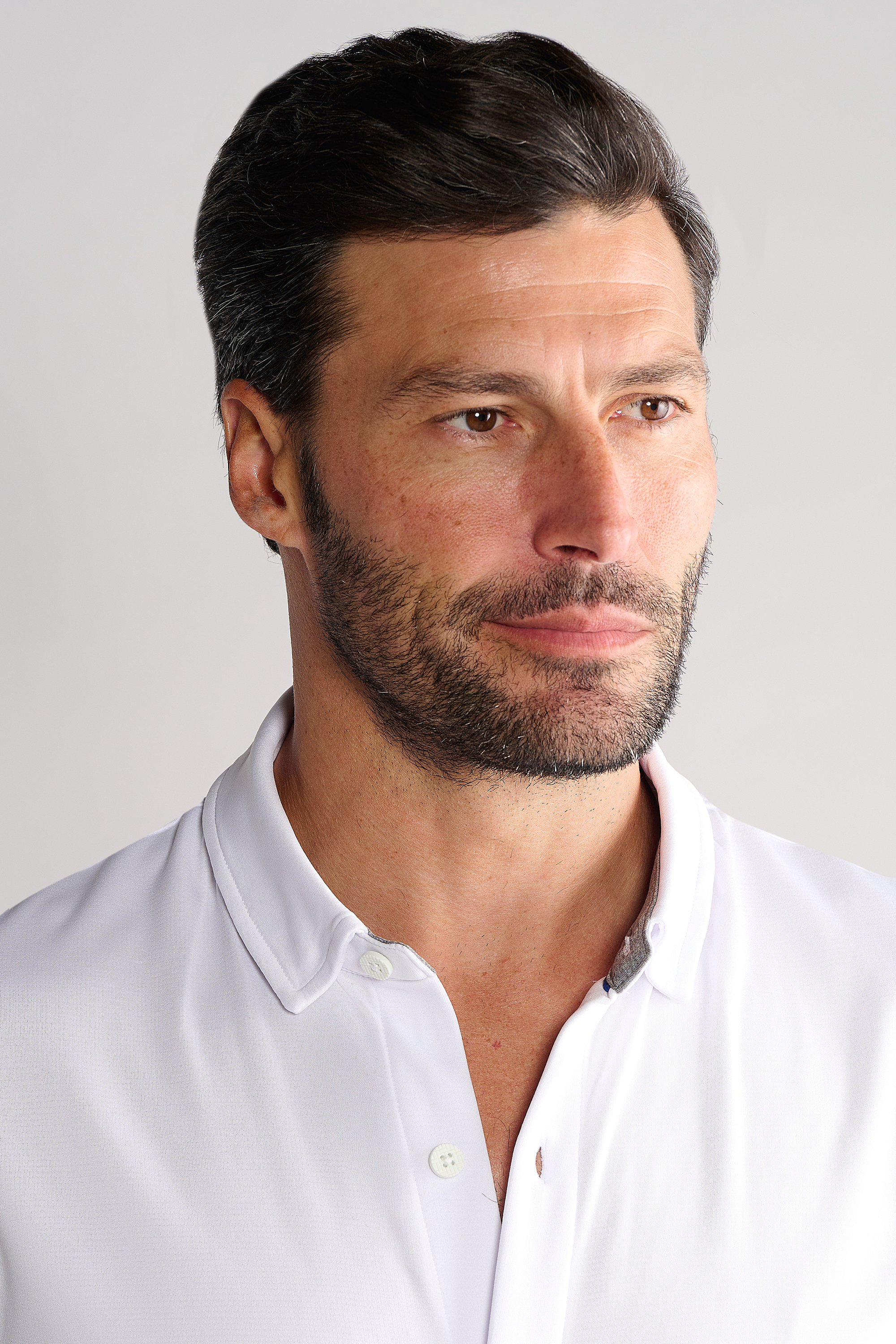 A man with short dark hair and a trimmed beard wears the Twillory Short Sleeve Button Down Polo Solid in White. He looks slightly right with a neutral expression, standing against a plain light background that highlights his shirt.