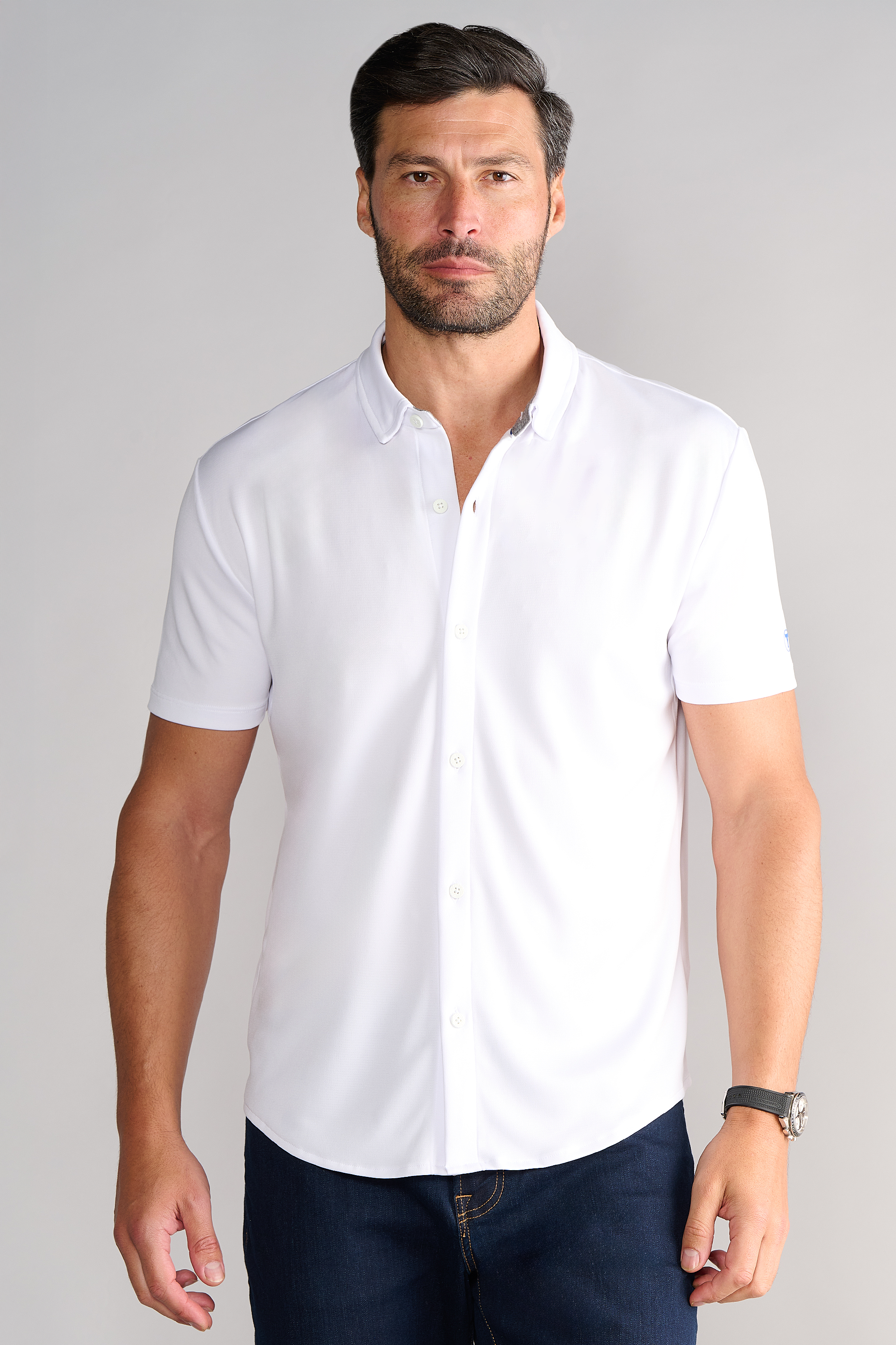 A man with short dark hair and a beard wears the Twillory Short Sleeve Button Down Polo Solid in white and dark jeans, standing facing forward against a plain light gray background with a neutral expression and a black watch.