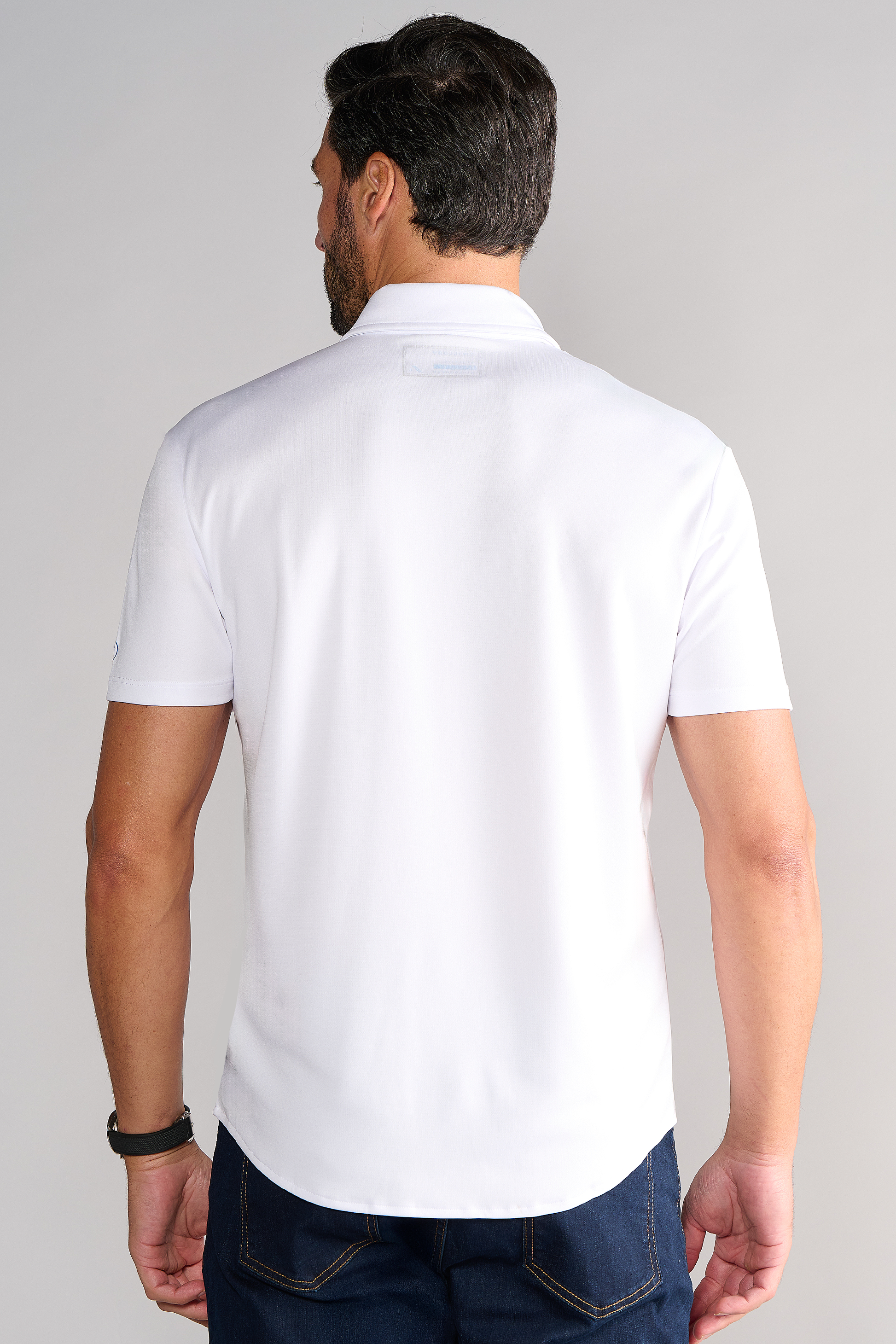 A bearded man with short dark hair faces away, wearing the Twillory Short Sleeve Button Down Polo Solid in White and dark blue jeans. He sports a black wristwatch on his left wrist, set against a plain light gray background.