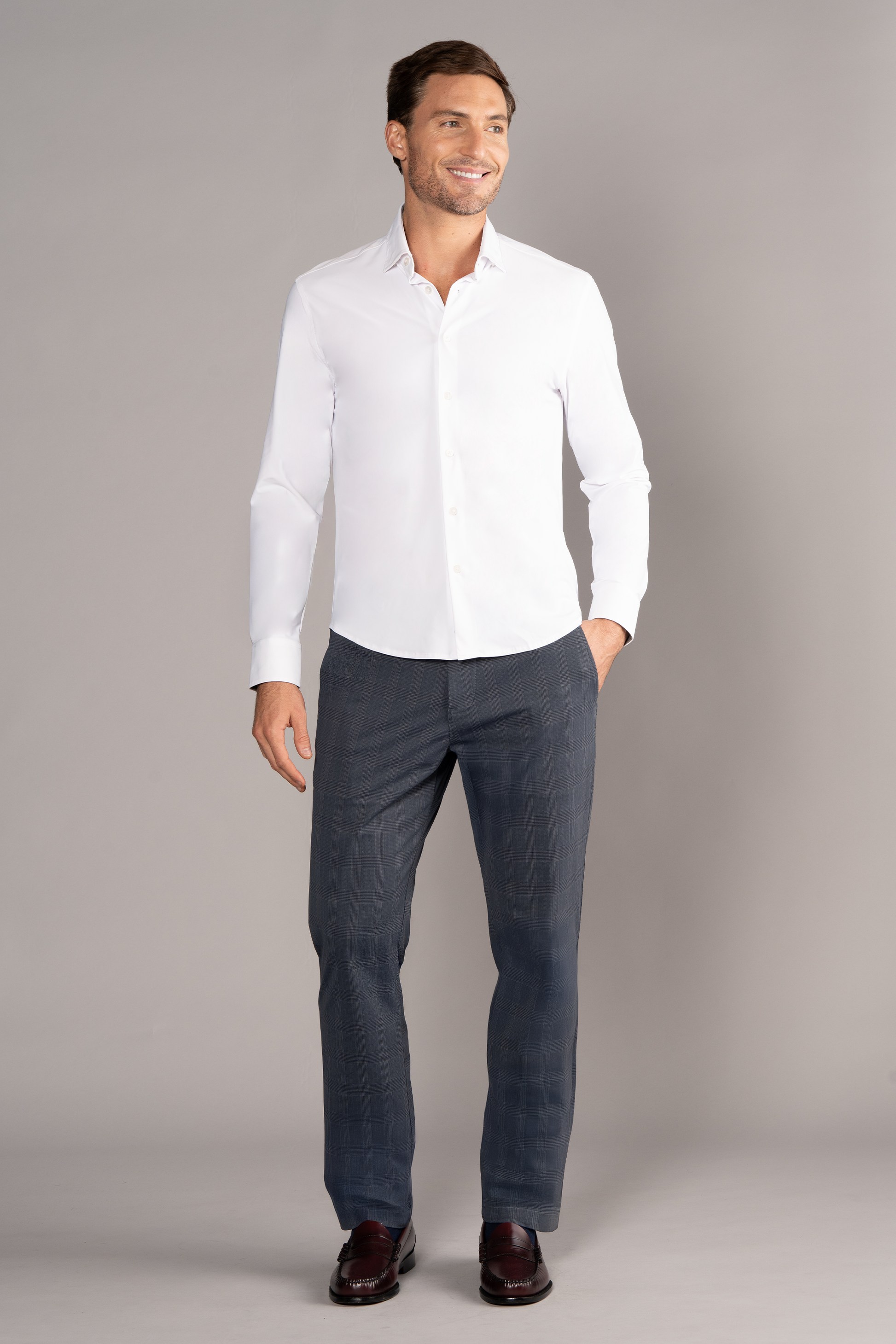A smiling man stands against a plain gray background, showcasing Twillory's The Event Shirt in white, paired with dark gray trousers and dark brown dress shoes. One hand is in his pocket as he looks slightly to the side.