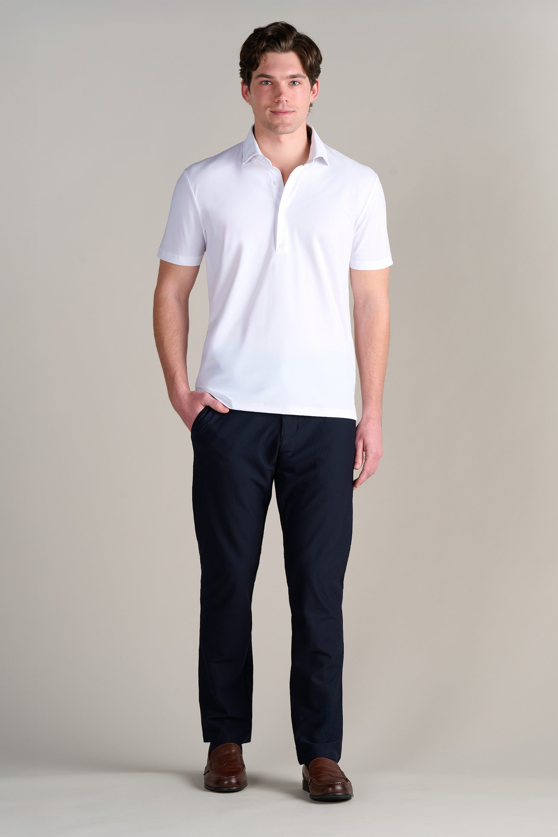 A man stands before a light-gray backdrop, wearing Twillory's Event Polo Solid in white, paired with dark navy trousers and brown dress shoes. One hand is in his pocket as he looks forward with a neutral expression.