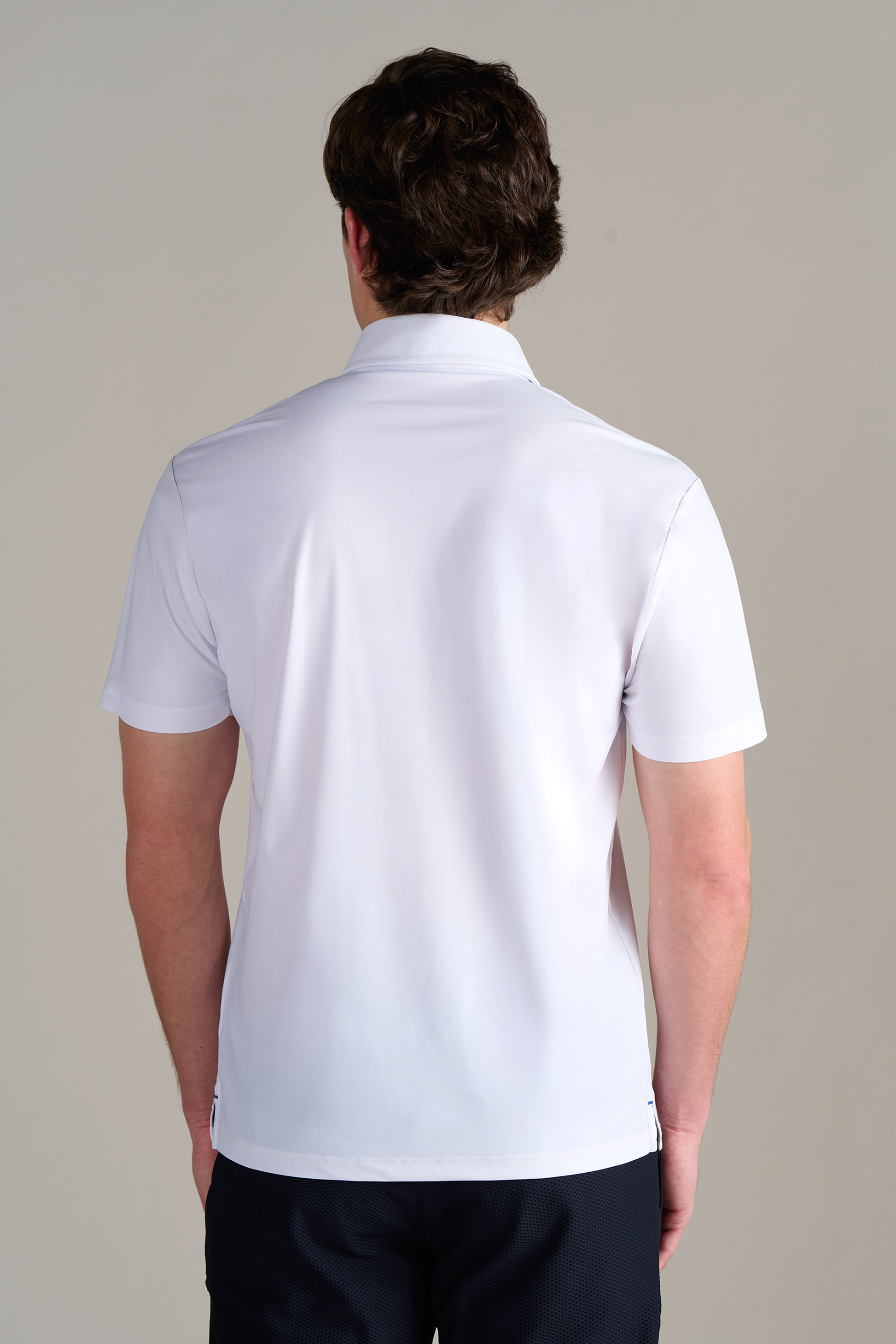 A man with short brown hair is seen from behind, wearing the Twillory Event Polo Solid in White. The shirt features a dress shirt collar and is wrinkle-free. He stands against a plain light grey background, arms relaxed at his sides.
