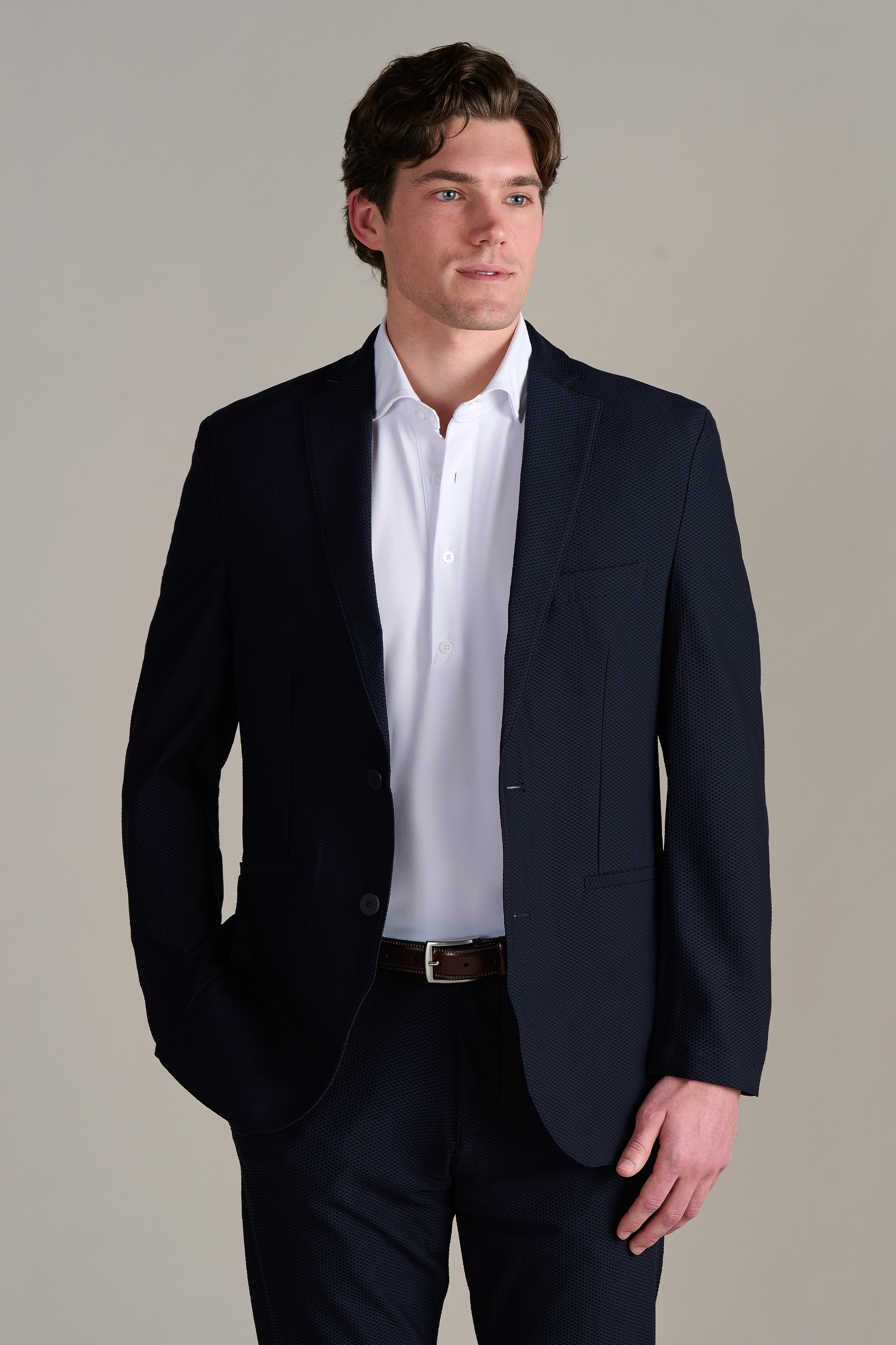 A man with wavy brown hair wears the Twillory Event Polo Solid in white, standing against a light gray background with one hand in his pocket and a neutral expression, looking slightly to his left.