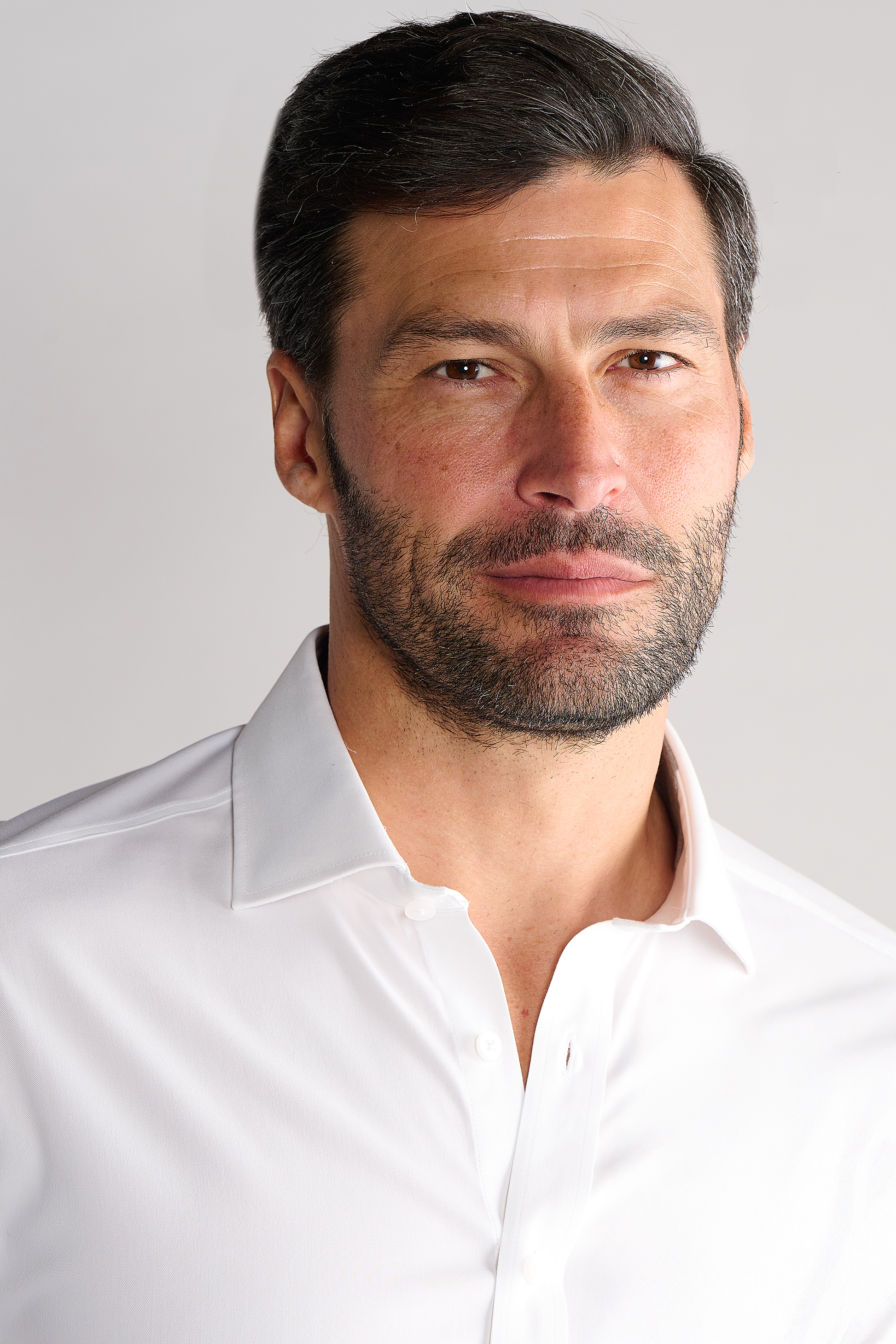 A man with short dark hair and a full beard wears the Twillory Leader Solid // White dress shirt. He faces the camera with a calm, confident expression and a slight smile, set against a plain light gray background.