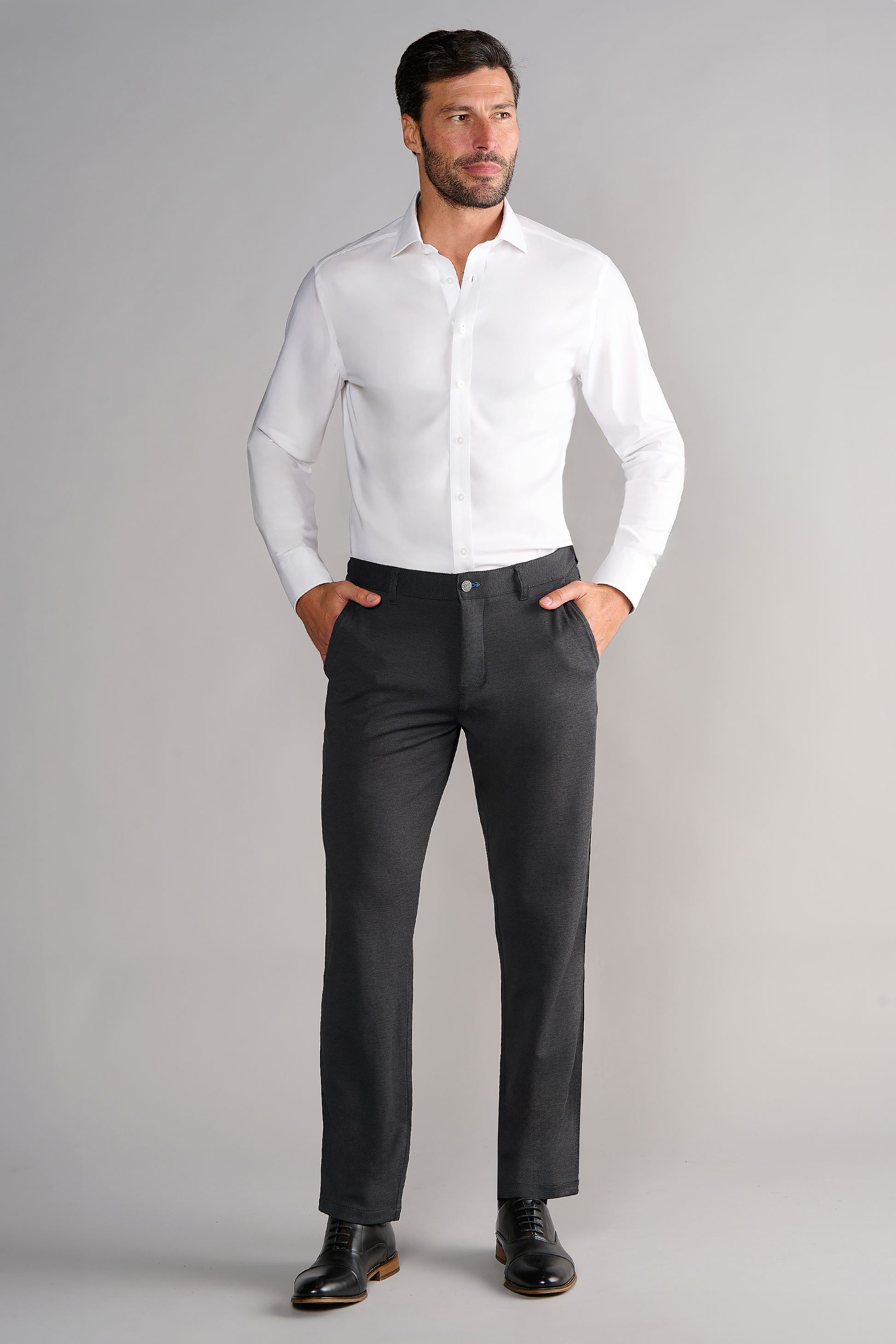 A man with short dark hair and a trimmed beard confidently poses against a light gray background wearing Twillory’s Leader Solid // White non-iron dress shirt, tucked into dark gray pants, paired with black dress shoes and hands in his pockets.