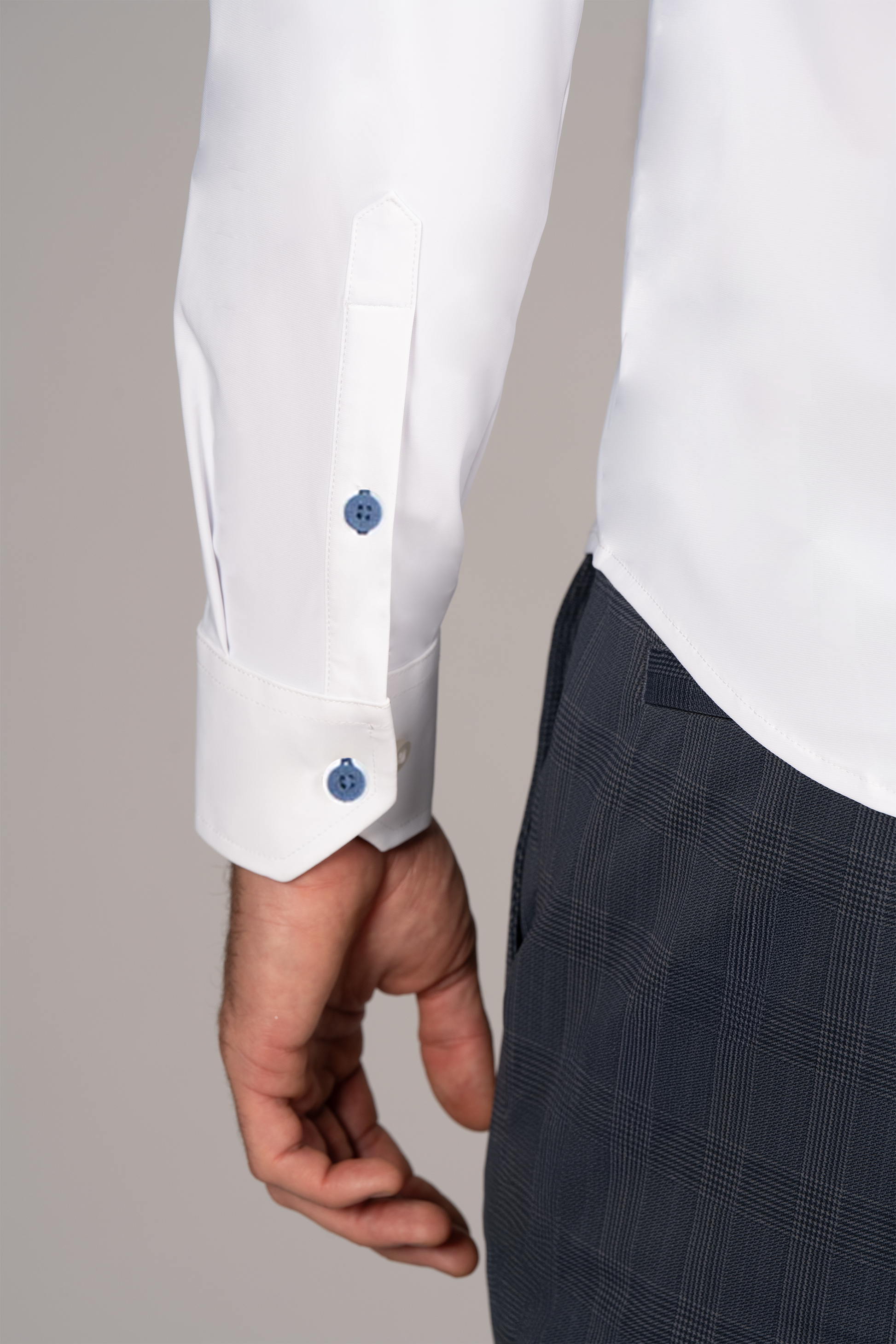 A person's left arm and hand are shown in The Event Shirt // White Blue by Twillory—stylish officewear with blue cuff buttons, made from wrinkle-free stretch, warp-knit tech fabric. The sleeve is buttoned, hand relaxed by their side, neutral background.