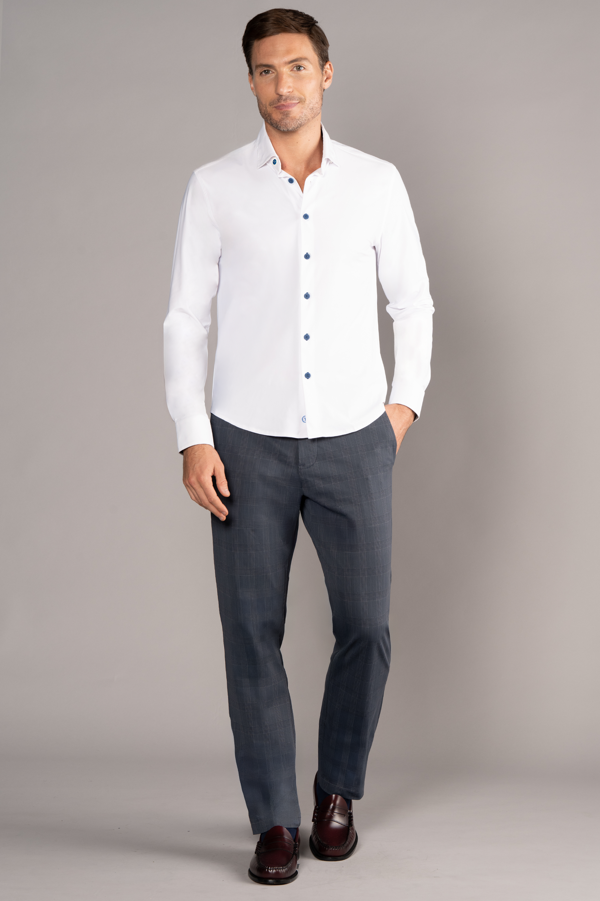 A man with short brown hair, a light beard, and a relaxed expression wears Twillory's The Event Shirt // White Blue against a plain gray background, paired with fitted gray plaid trousers and dark brown dress shoes.