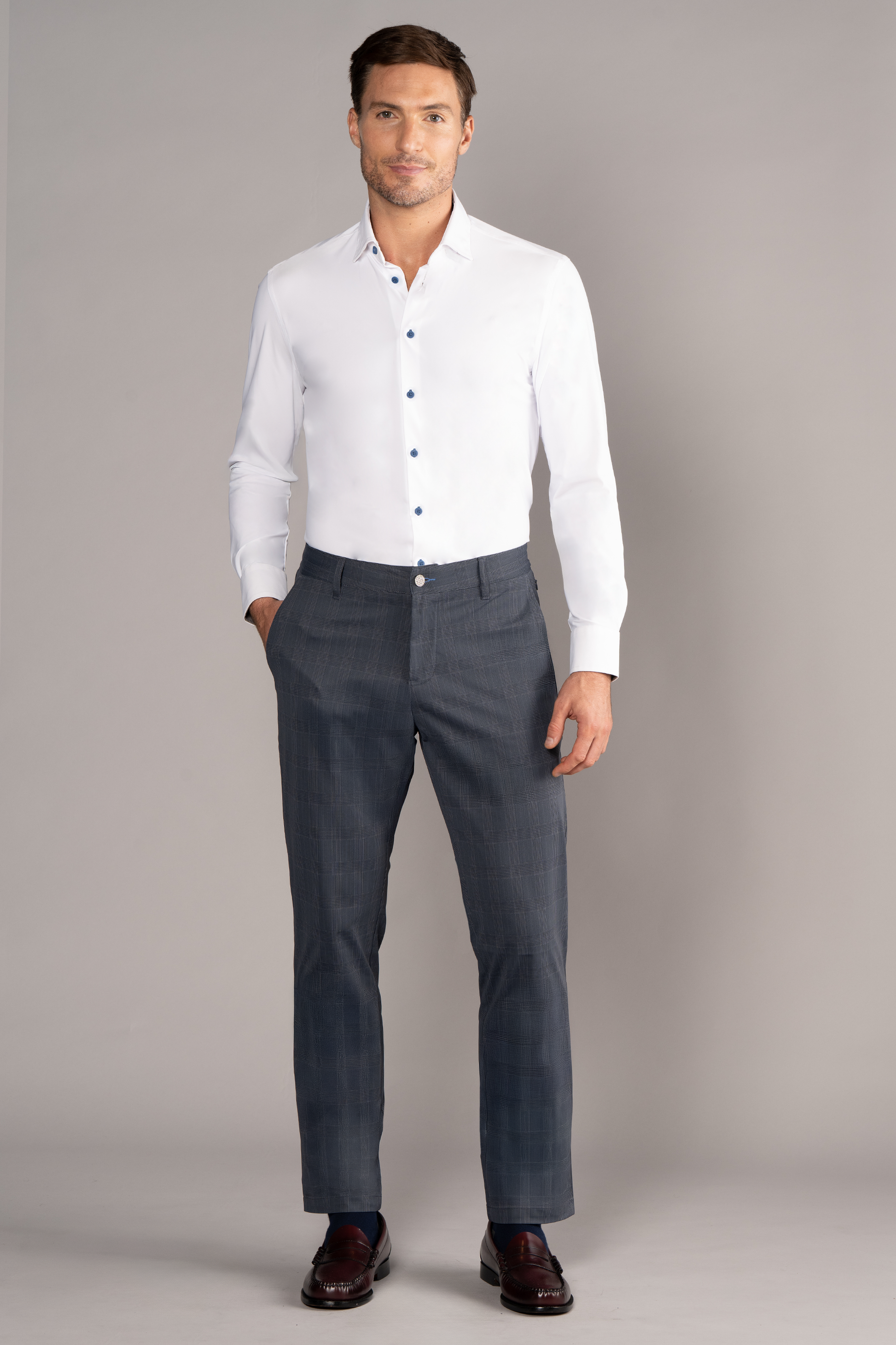 A man stands facing forward in The Event Shirt // White Blue by Twillory—a fitted, wrinkle-free stretch white dress shirt with blue buttons—paired with gray dress pants and dark brown loafers. One hand is in his pocket, making for perfect officewear.