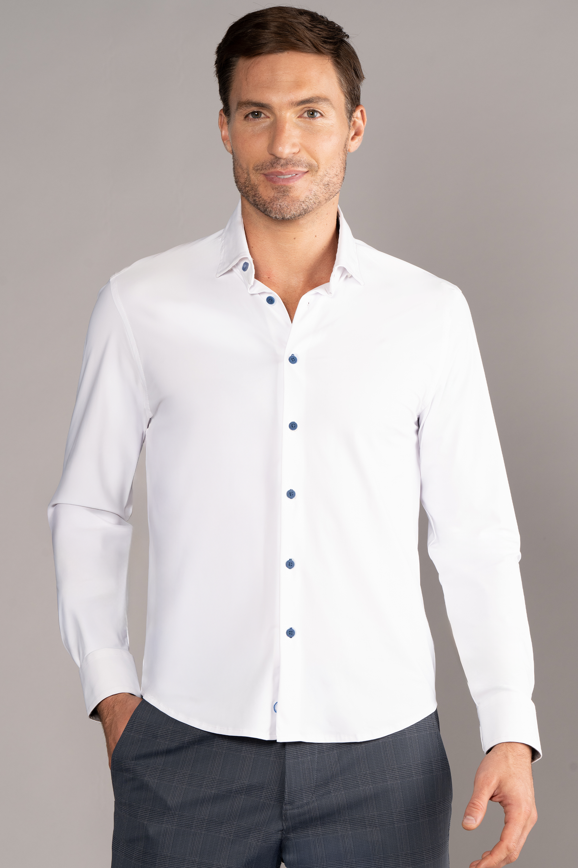 A man with short brown hair and light skin smiles in front of a neutral background, wearing Twillory’s The Event Shirt // White Blue—a fitted, wrinkle-free stretch button-up with blue buttons—paired with gray dress pants.