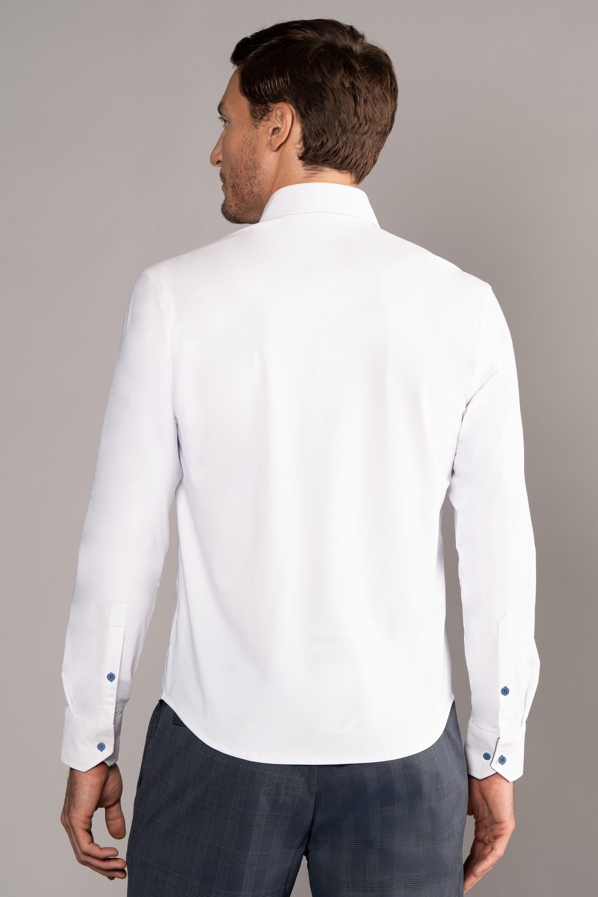 A man with short brown hair is seen from behind wearing Twillory's The Event Shirt // White Blue— a wrinkle-free, stretch white dress shirt with blue buttons, paired with gray plaid trousers. Perfect for officewear. Background is plain light gray.