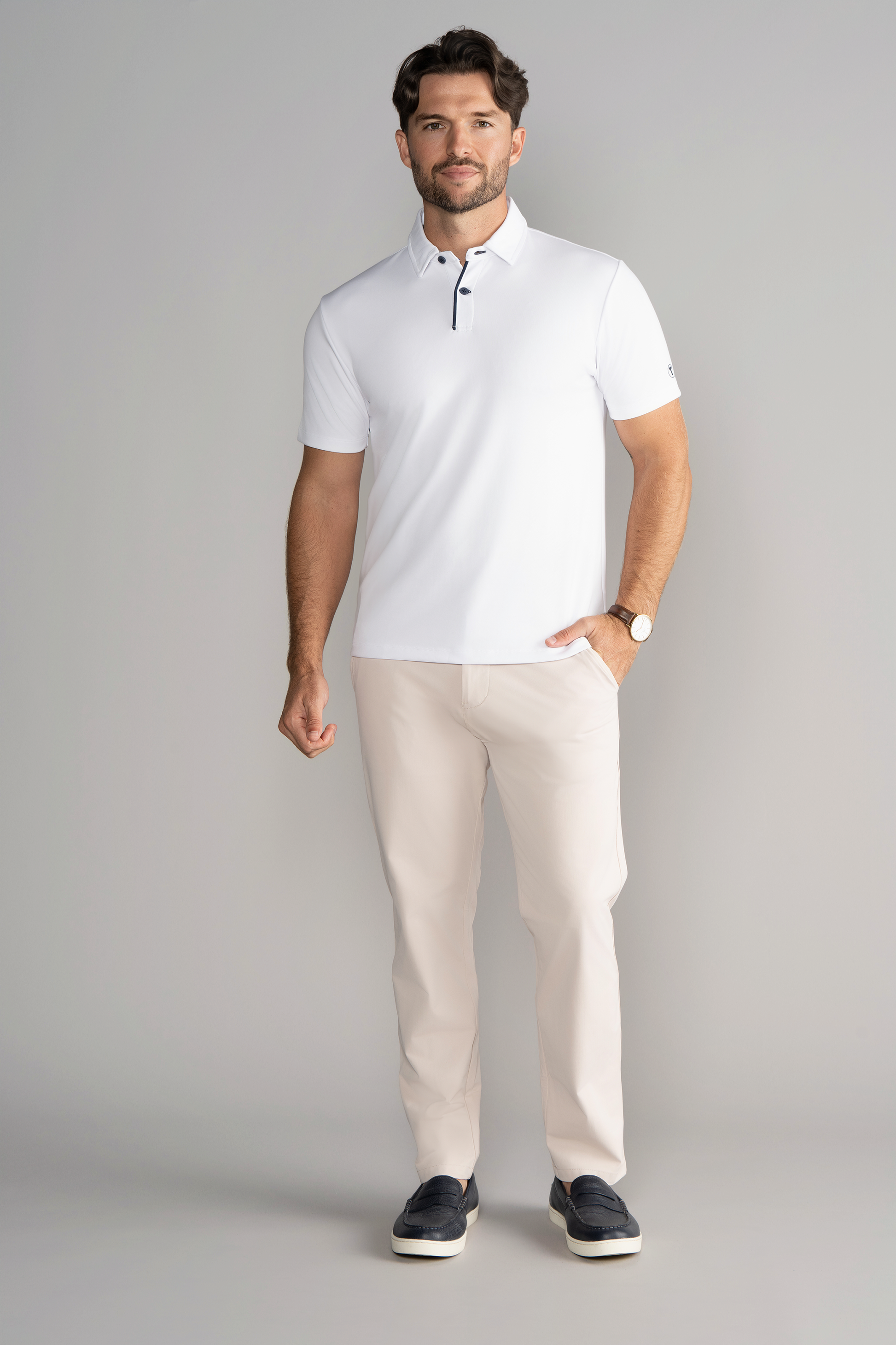 A man with short brown hair and a beard stands against a gray background, wearing Twillory's Short Sleeve Polo Contrast in White Navy, light beige pants, dark slip-on shoes, and a wristwatch. He smiles slightly with one hand in his pocket.