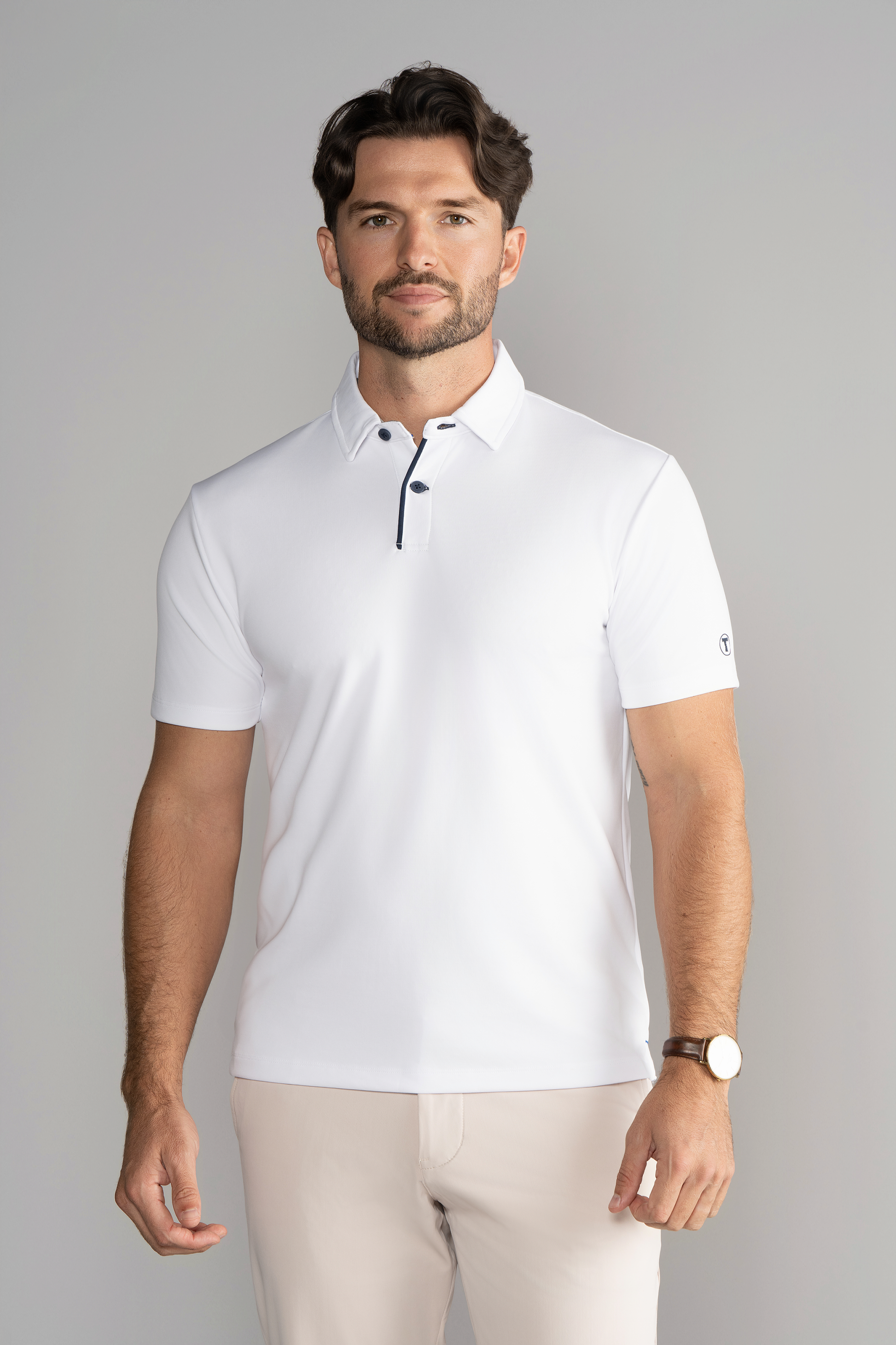 A man with short brown hair and a trimmed beard wears the Twillory Short Sleeve Polo Contrast in White Navy and light beige pants. He faces forward with a neutral expression, wearing a brown watch, against a light gray background.