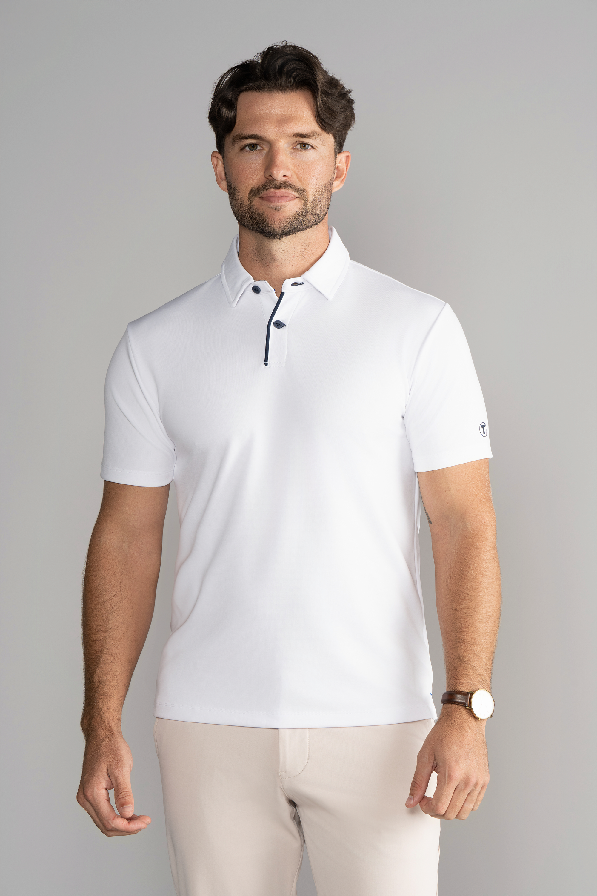 A man with short brown hair and a trimmed beard wears the Twillory Short Sleeve Polo Contrast in White Navy and light beige pants. He faces forward with a neutral expression, wearing a brown watch, against a light gray background.