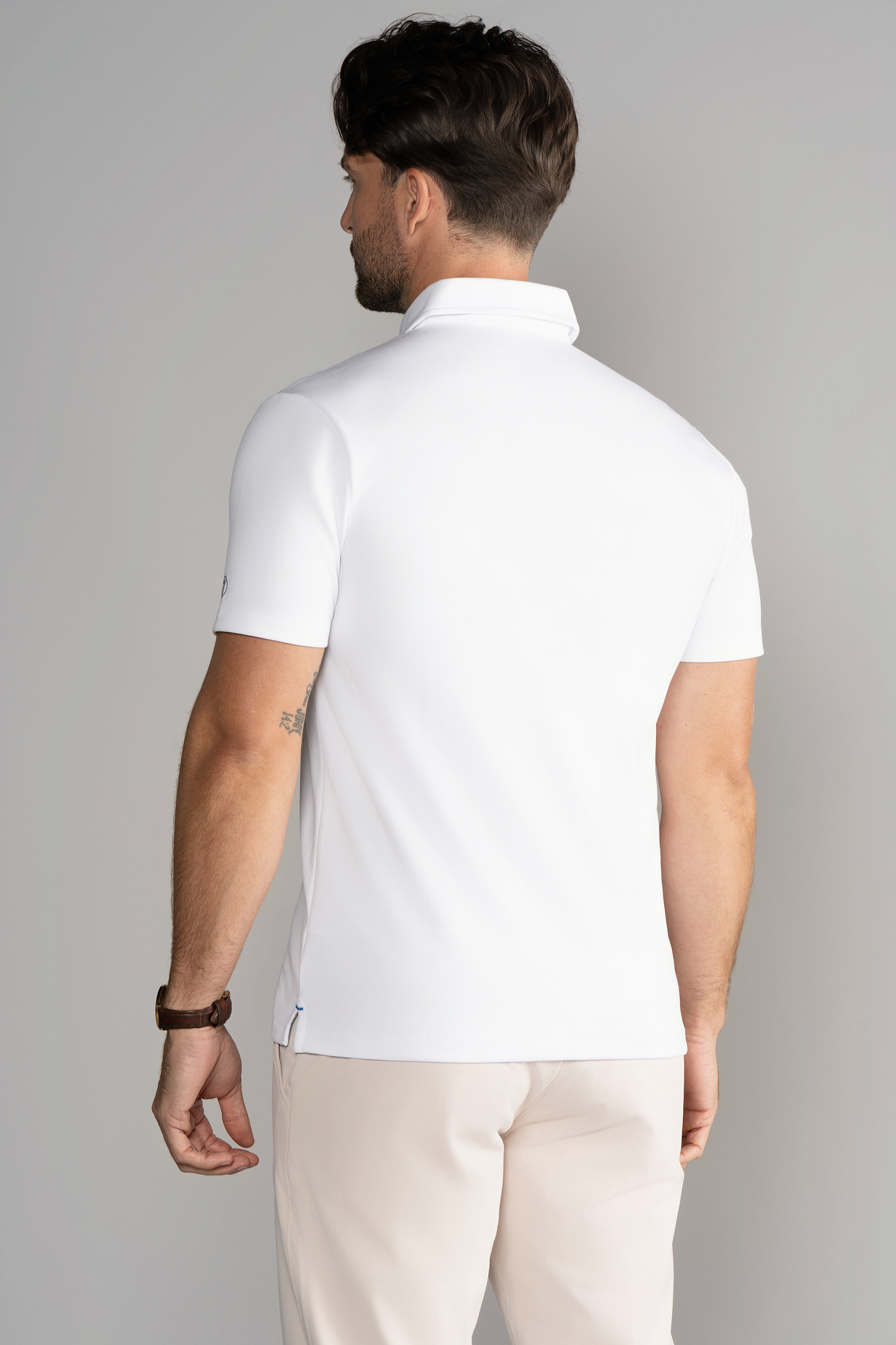 A man with short brown hair is seen from behind wearing the Twillory Short Sleeve Polo Contrast in White Navy and light beige pants. He has a brown watch on his left wrist, a small tattoo on his right upper arm, and stands against a light gray background.