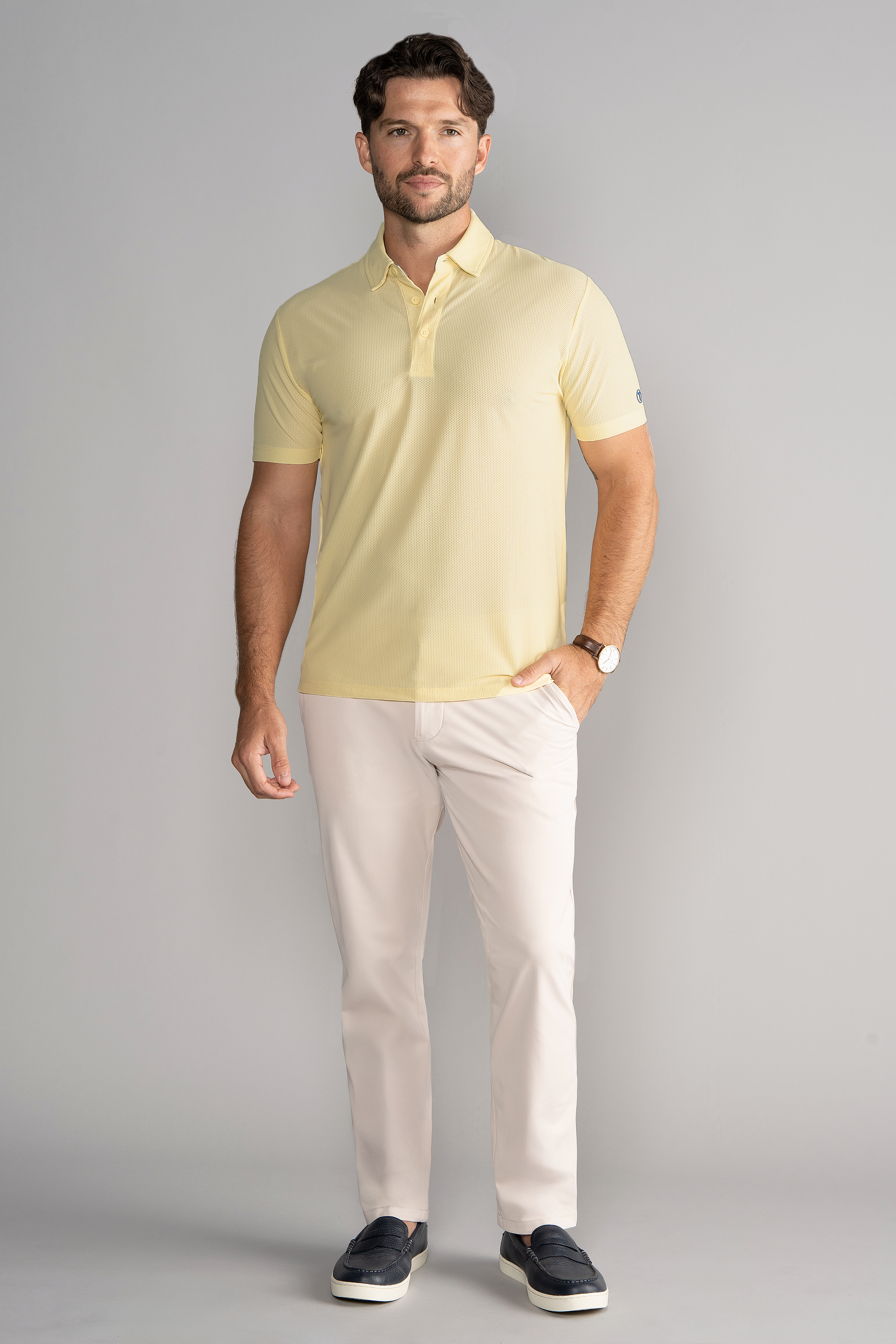 A man with short brown hair and a trimmed beard poses casually against a light gray background, wearing Twillory’s AIR 3 Button Polo Solid in yellow, paired with light beige pants and dark navy loafers.