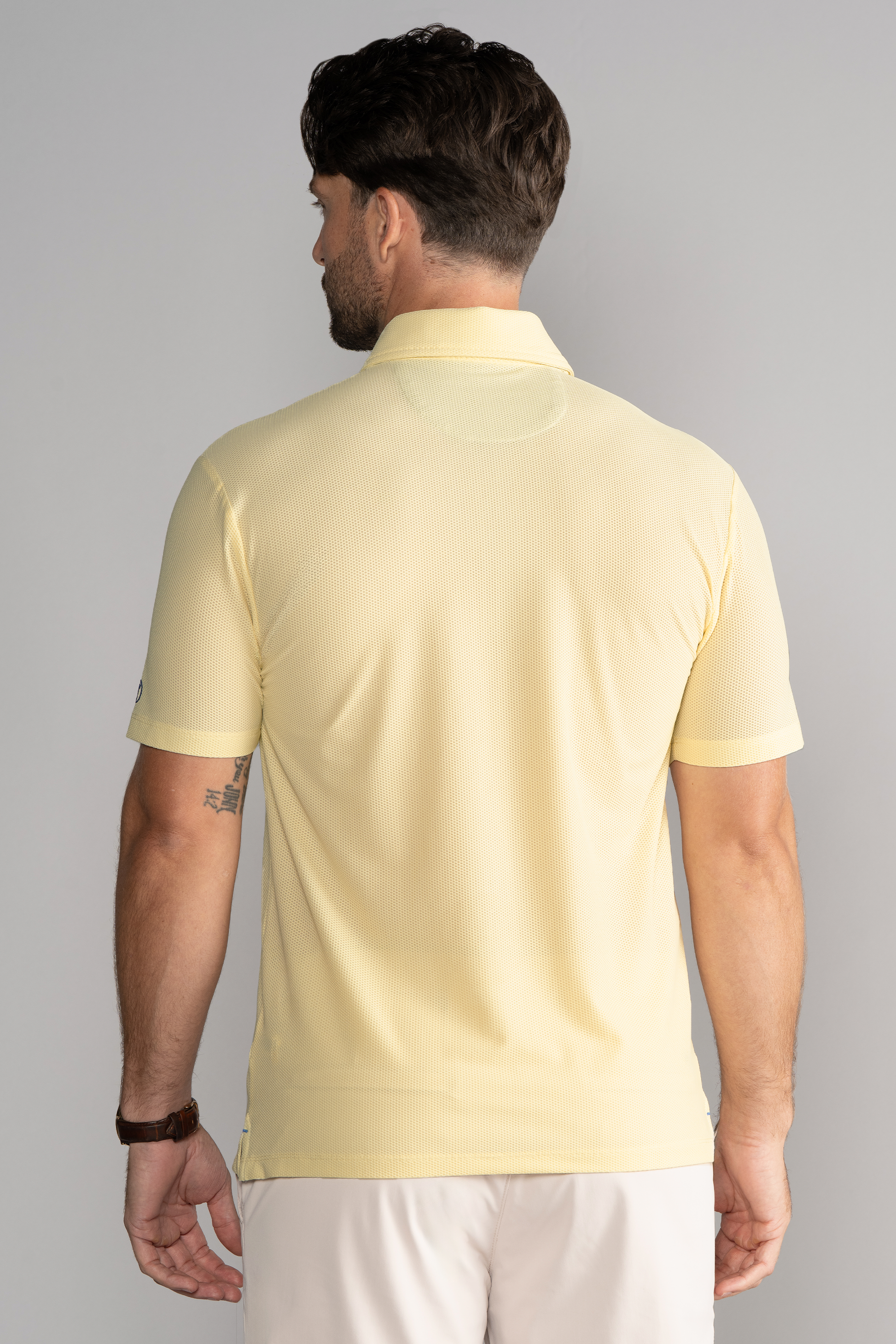 A man with short dark hair, shown from behind, wears a Twillory AIR 3 Button Polo Solid in yellow and light shorts. He has a right upper arm tattoo, a brown watch on his left wrist, and stands against a plain gray background.