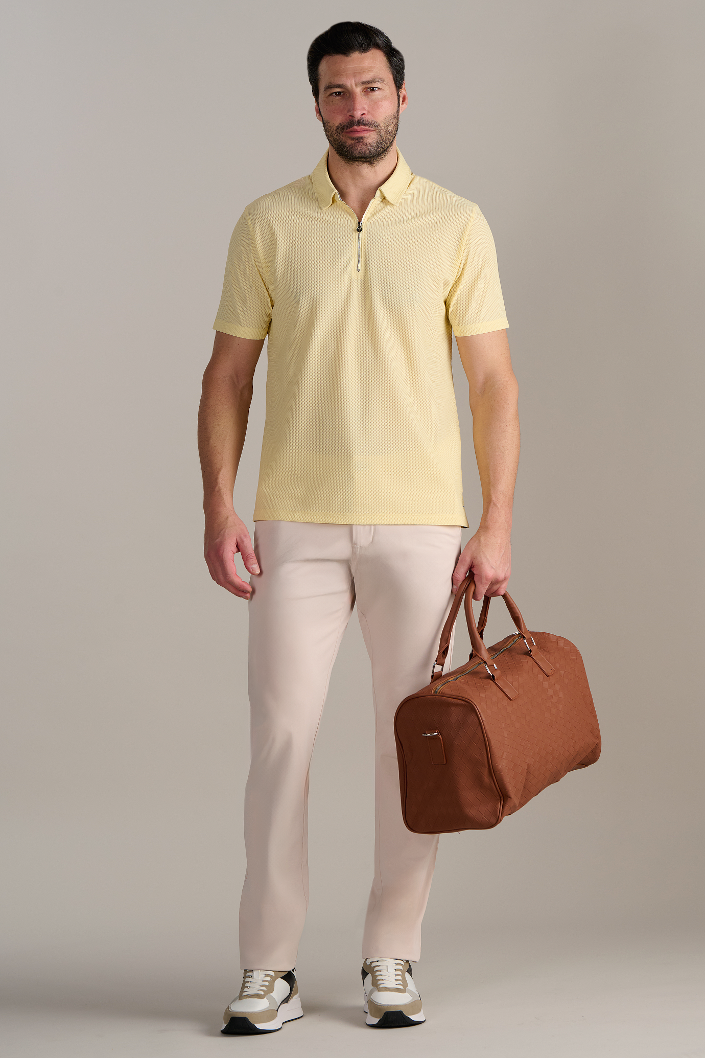 A man with short dark hair and a beard faces forward, wearing Twillory's AIR Zip Polo Solid in yellow, light beige pants, white and beige sneakers, and holding a brown duffel bag against a plain light gray background.