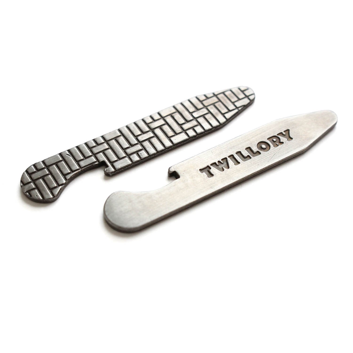 Two silver-colored Bottle Opener Stays by Twillory are shown on a white background. One features a woven pattern, the other is engraved with TWILLORY. Both have side notches and rounded ends, designed to pair perfectly with Twillory shirts.