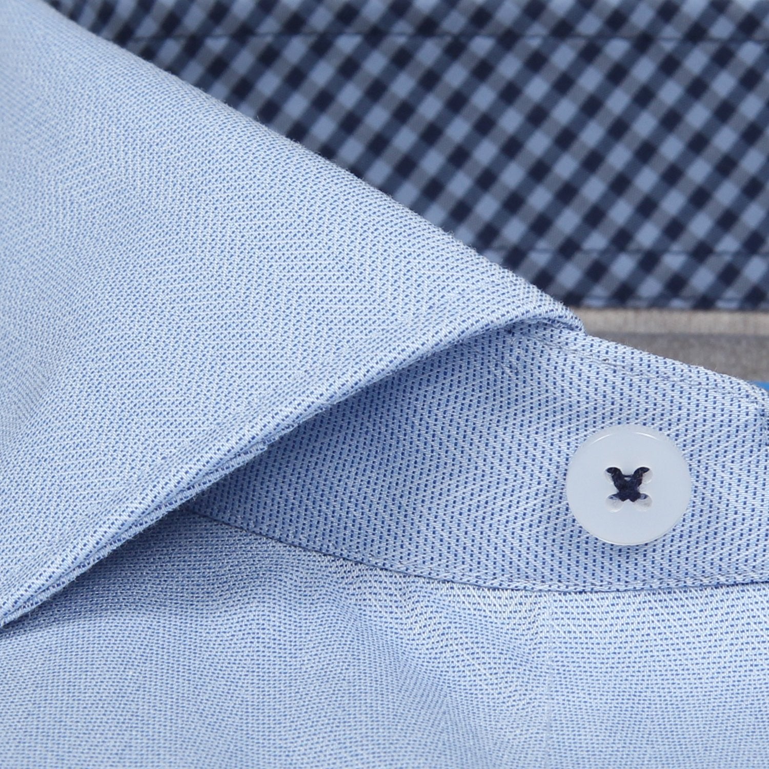 Close-up of a light blue dress shirt collar with a button detail and blue checkered fabric background.