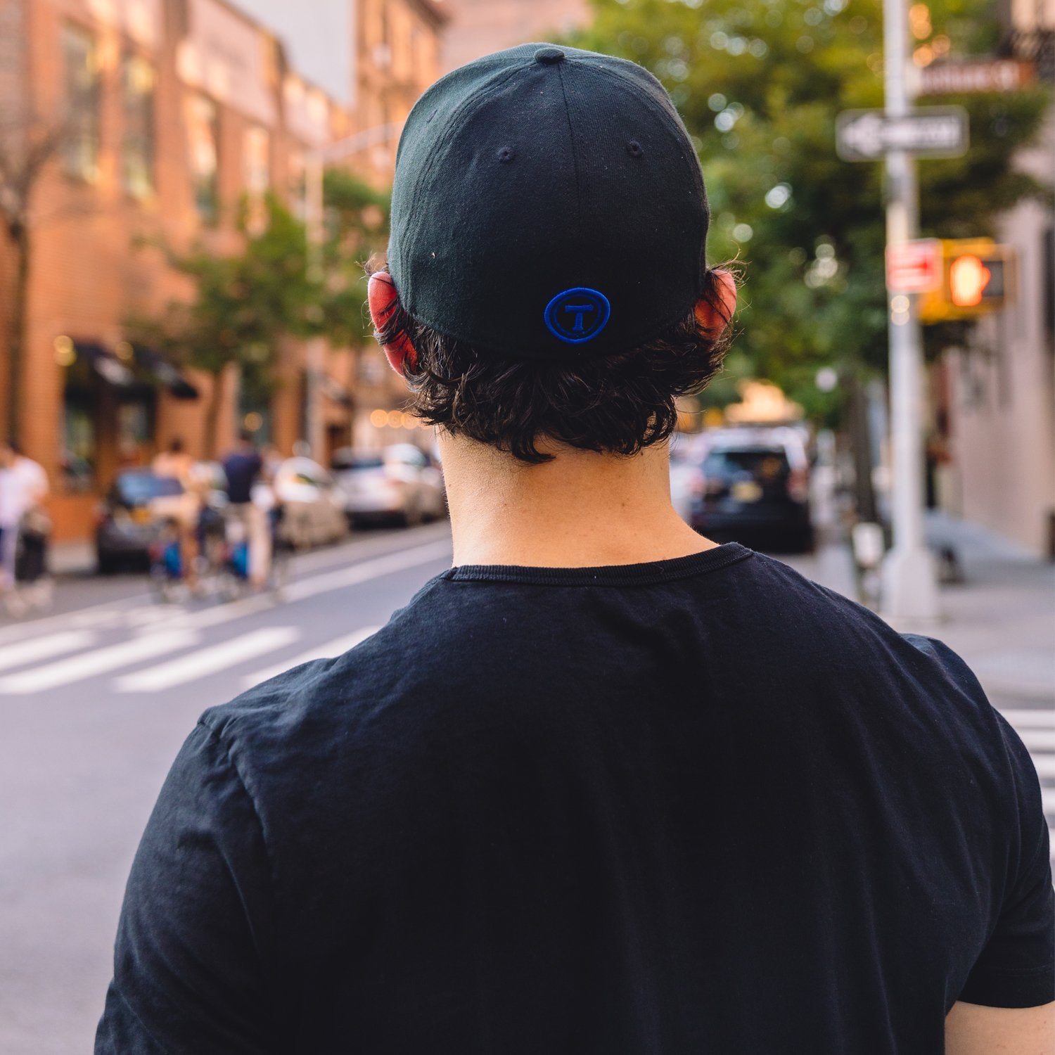 A person wearing a black T-shirt and a Twillory Twill Cap featuring a blue circular logo stands with their back to the camera on a sunlit, bustling city street lined with buildings, trees, and parked cars.