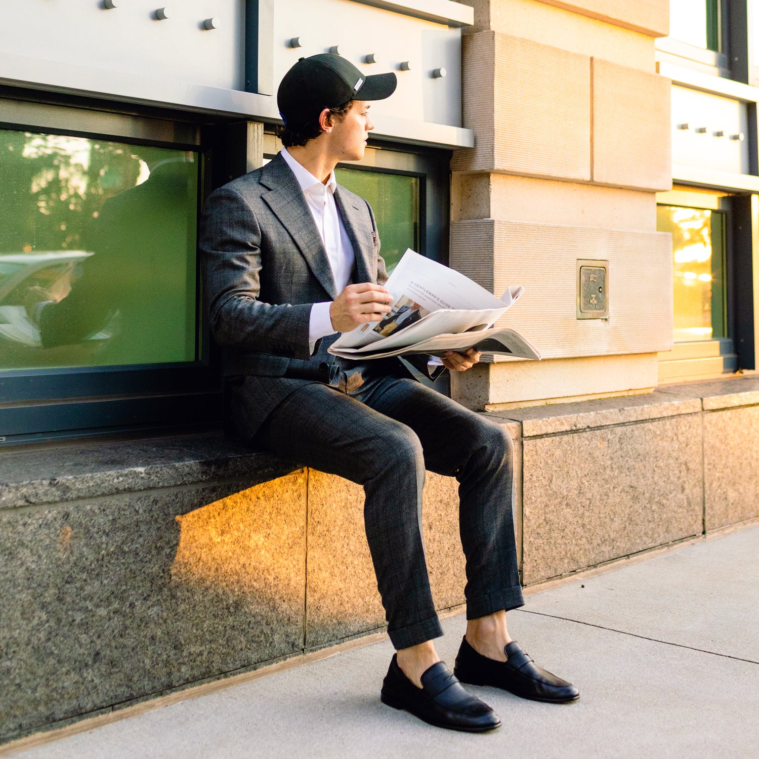 A man in a dark suit and M-L size Twillory Twill Cap sits on a stone ledge outside, holding an open newspaper. He's wearing black loafers without socks and gazes left as warm sunlight washes over the scene.