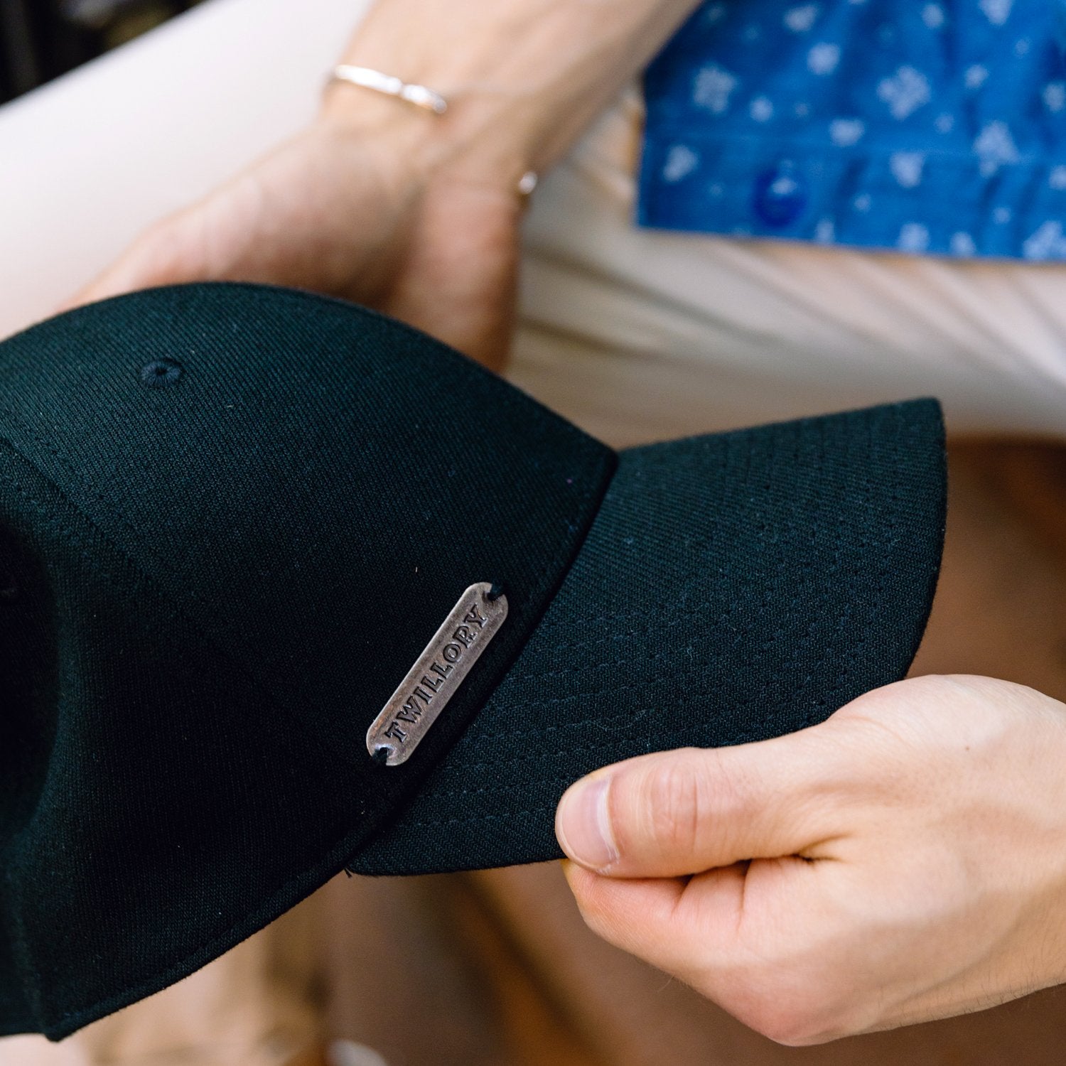 A person holds the Twillory Twill Cap—a black baseball cap with a metal plaque—in both hands. Dressed in light pants, a blue patterned shirt, and a silver bracelet, they pose before a blurred background.