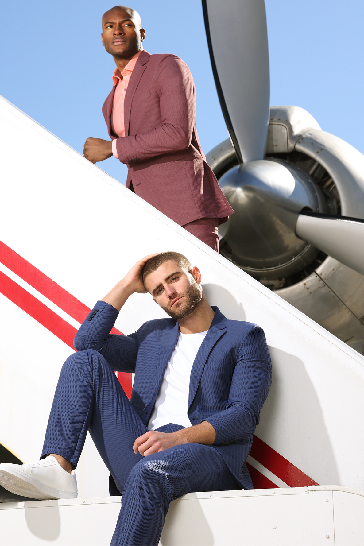 Two men in stylish suits posing on an airplane staircase under a clear blue sky; one in a burgundy suit standing and looking off into the distance, the other in a blue suit sitting with a relaxed pose.