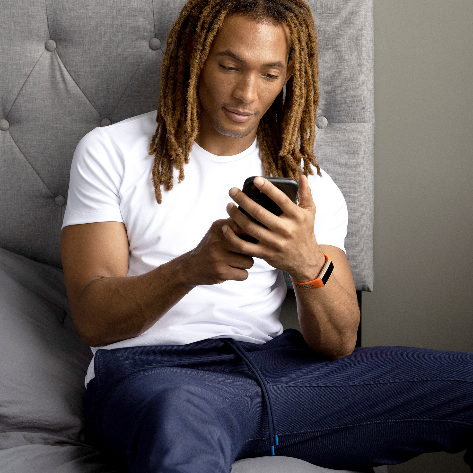Man with dreadlocks using a smartphone while sitting on a bed, wearing a white t-shirt and blue pants.