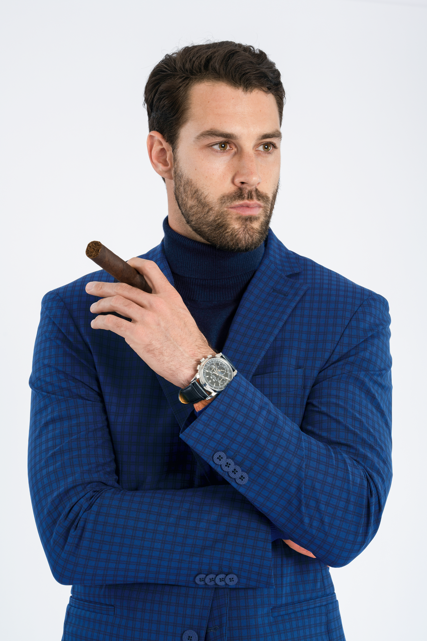A bearded man wearing Twillory's Performance Blazer in Blue Check and a dark turtleneck stands against a white background, holding an unlit cigar with his left hand across his chest and sporting a large silver watch, gazing thoughtfully to the side.