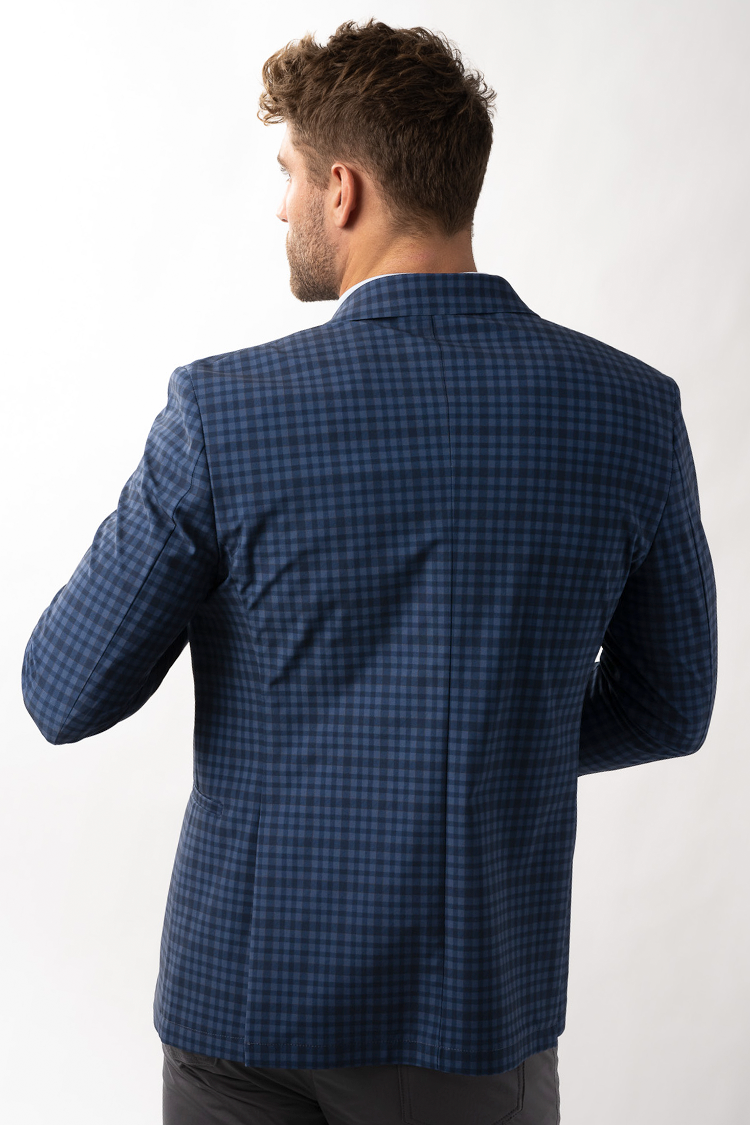 A man with short, curly brown hair, shown from behind against a white background, wears the Twillory Performance Blazer in Blue Navy Check. His right arm is bent and his face is partly in profile.