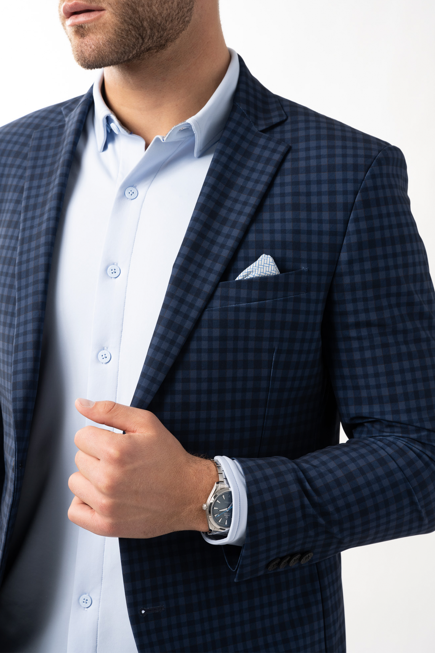 A man with light stubble wears the Twillory Performance Blazer // Blue Navy Check over a light blue shirt. He holds the lapel, highlighting 3D shoulder pads and a pocket square, against a white background.