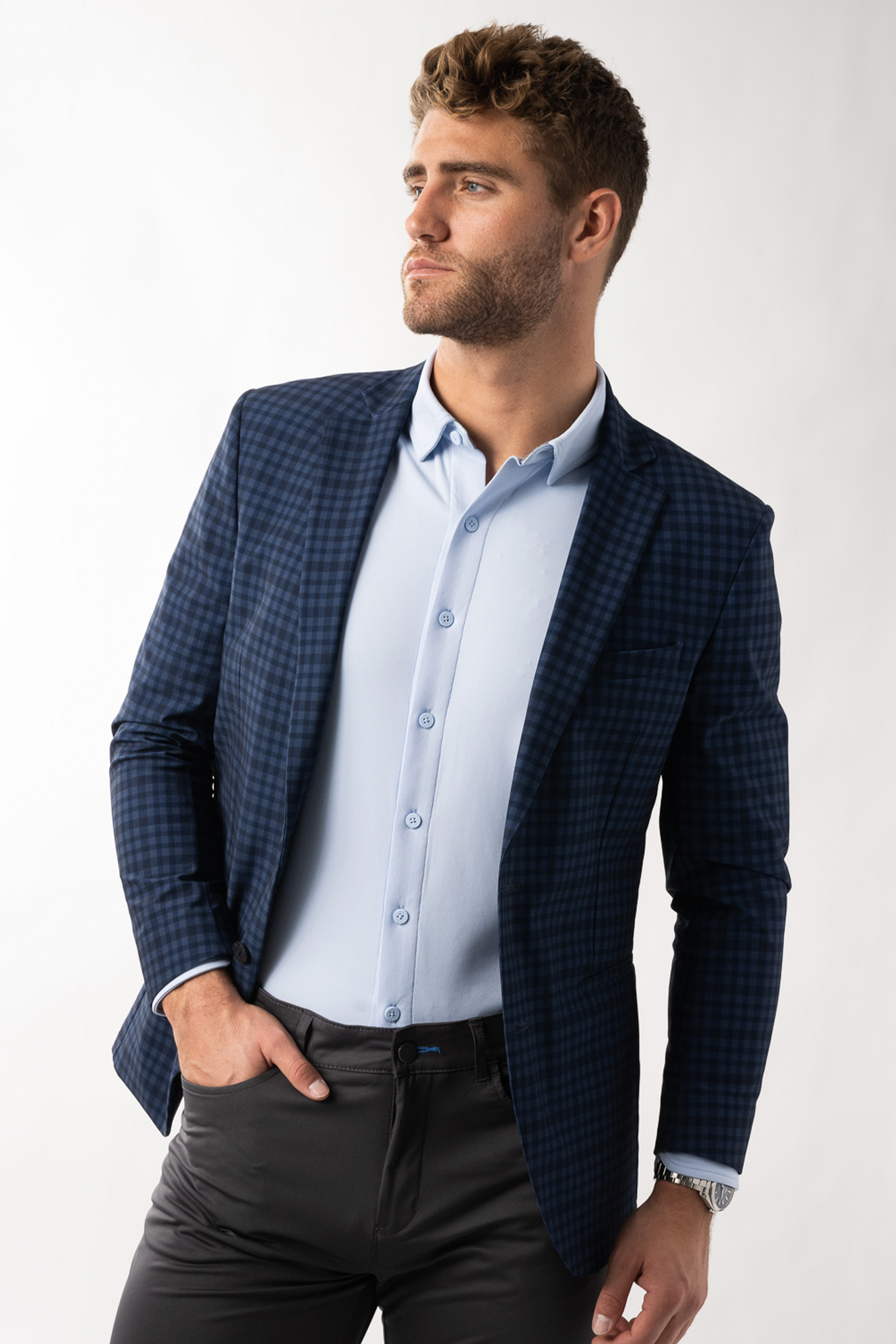 A man with short, curly brown hair and a beard wears the Twillory Performance Blazer in Blue Navy Check over a light blue shirt and dark slacks, looking left with one hand in his pocket against a plain, light background.
