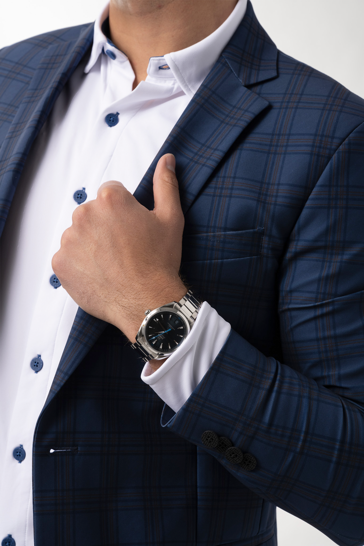 A person in Twillory’s Performance Blazer // Blue Plaid, made from Japanese stretch fabric with subtle red lines and blue buttons, holds their lapel to reveal a silver wristwatch against a plain white background.
