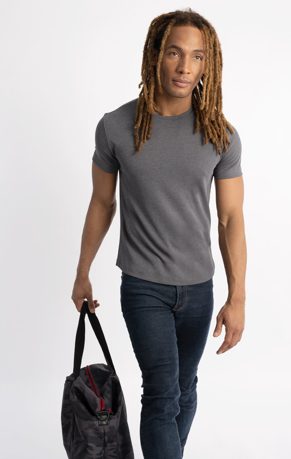 Man with long dreadlocks wearing a gray t-shirt and jeans, carrying a camouflage-patterned duffle bag, walking against a plain white background.