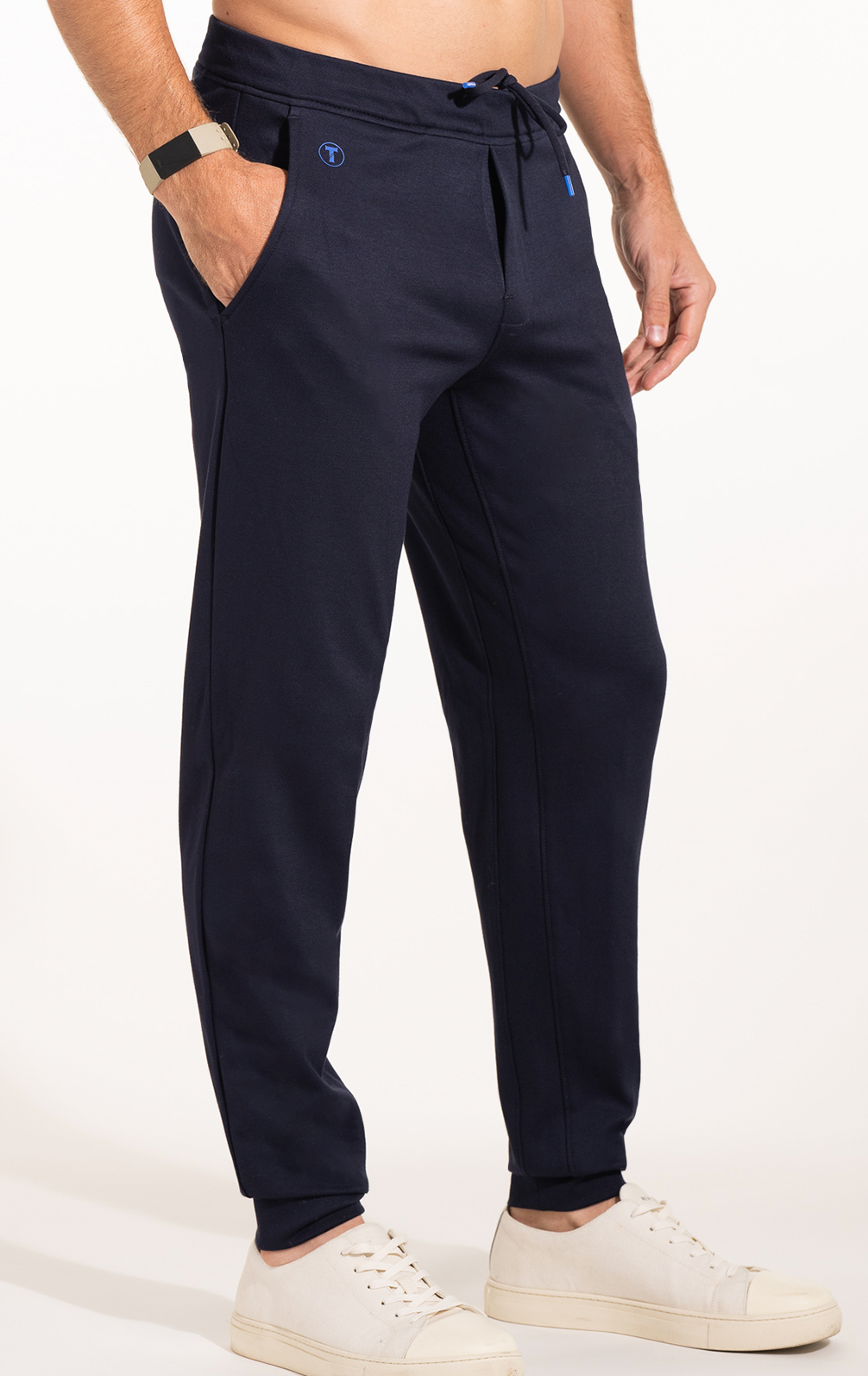 A man wears Twillory Athlounger Jogger pants in navy with a tapered fit and drawstring waist, paired with a black smartwatch and white low-top sneakers. The background is plain white; his torso is partially out of frame.
