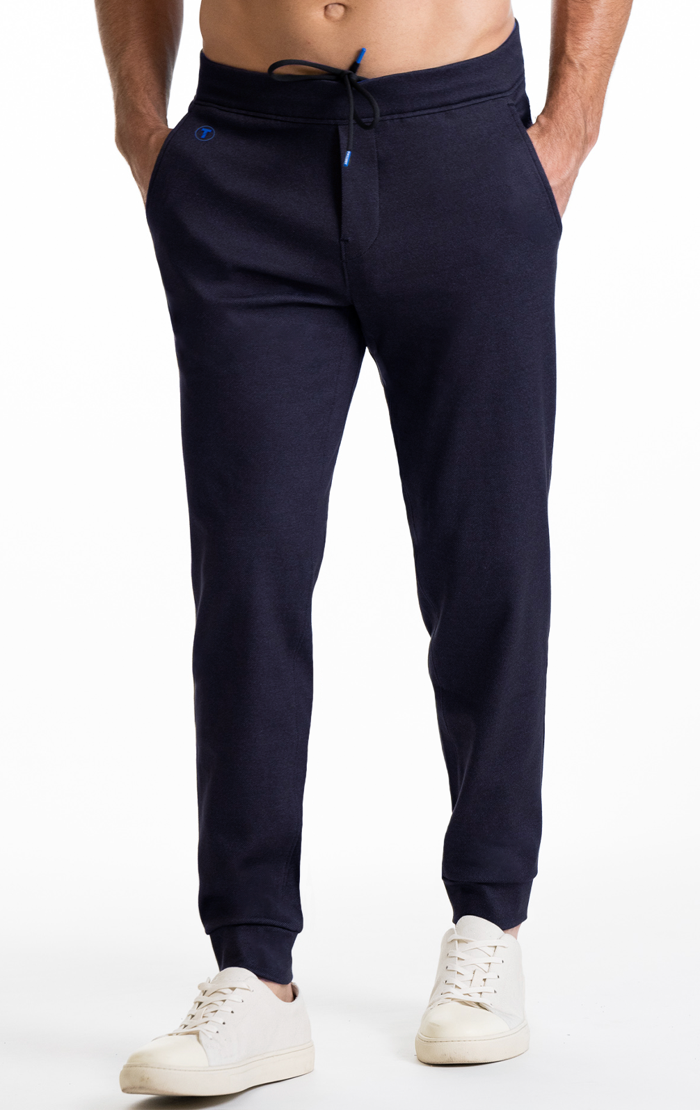 A person wears Twillory Athlounger Joggers in dark navy, featuring comfort fabric, a drawstring waist, and a side-zip pocket, paired with white low-top sneakers. The image shows them from mid-torso down against a white background.