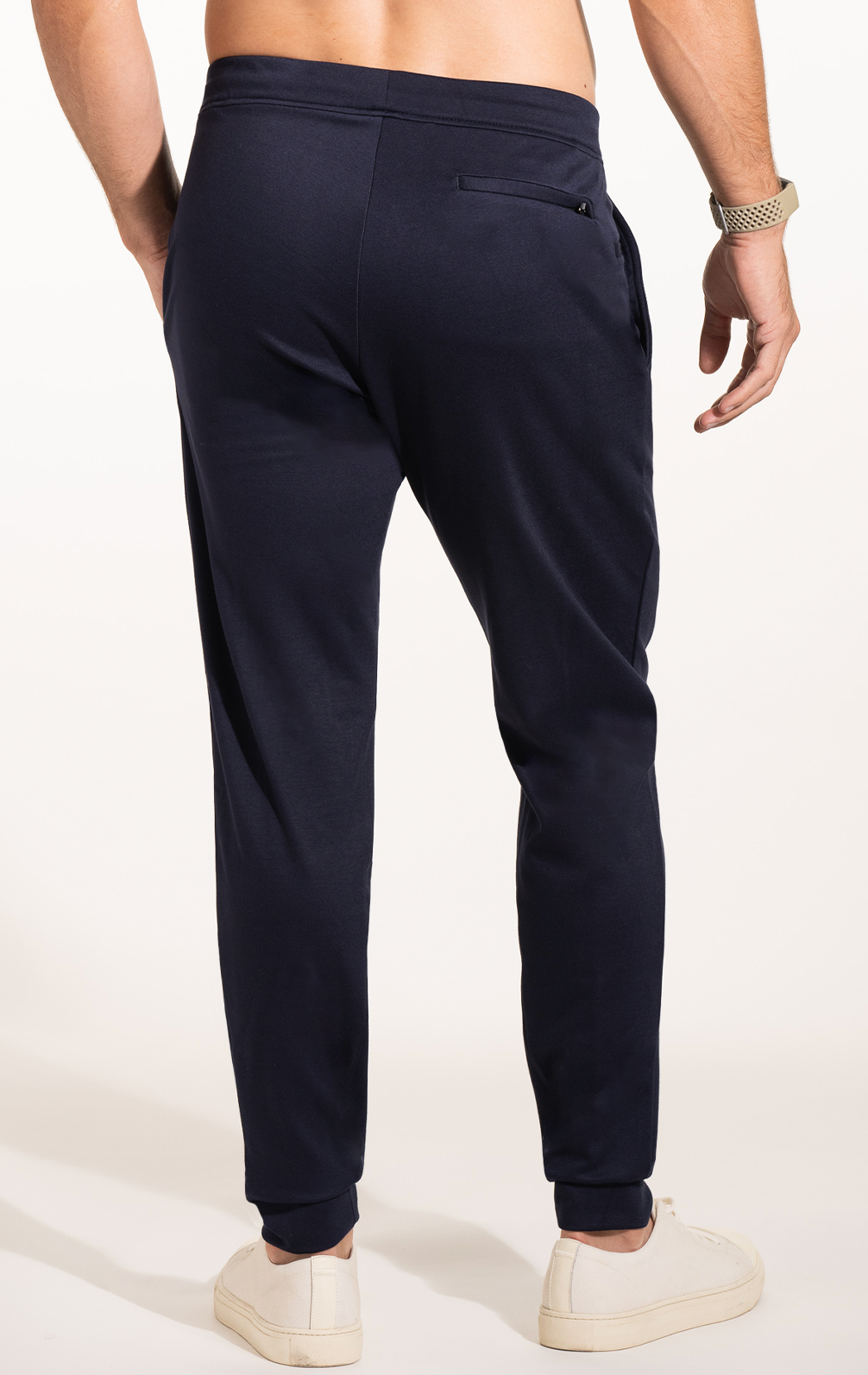 A man, shirtless and wearing a watch, models Twillory’s Athlounger Jogger in navy blue. The comfort fabric joggers feature a right side-zip pocket. He pairs them with white sneakers against a plain, well-lit background.