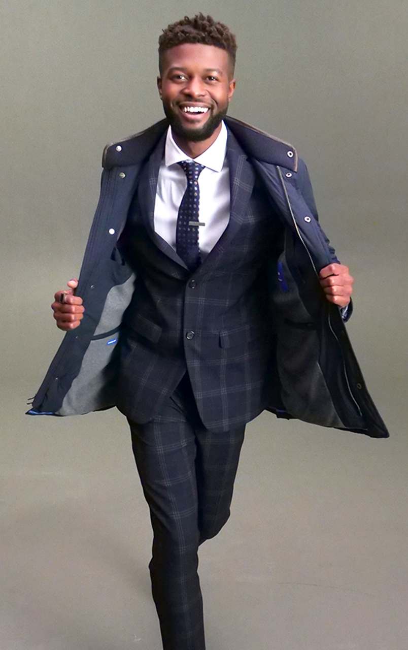 Man confidently walking in a stylish dark plaid suit with coat open, smiling and looking directly at the camera.