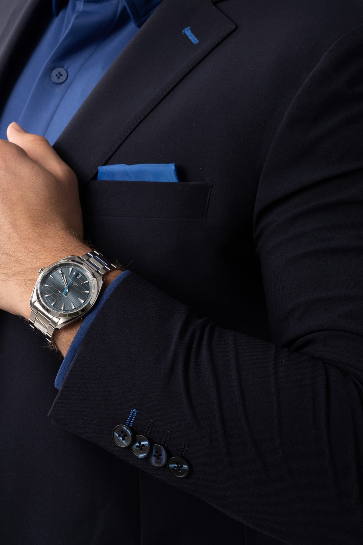 A close-up of a person wearing Twillory's Performance Blazer // NAVY BLUE CONTRAST, a blue dress shirt, and blue pocket square. Their left hand touches their chest, showing a silver watch with a blue face. Four blazer sleeve buttons are visible.