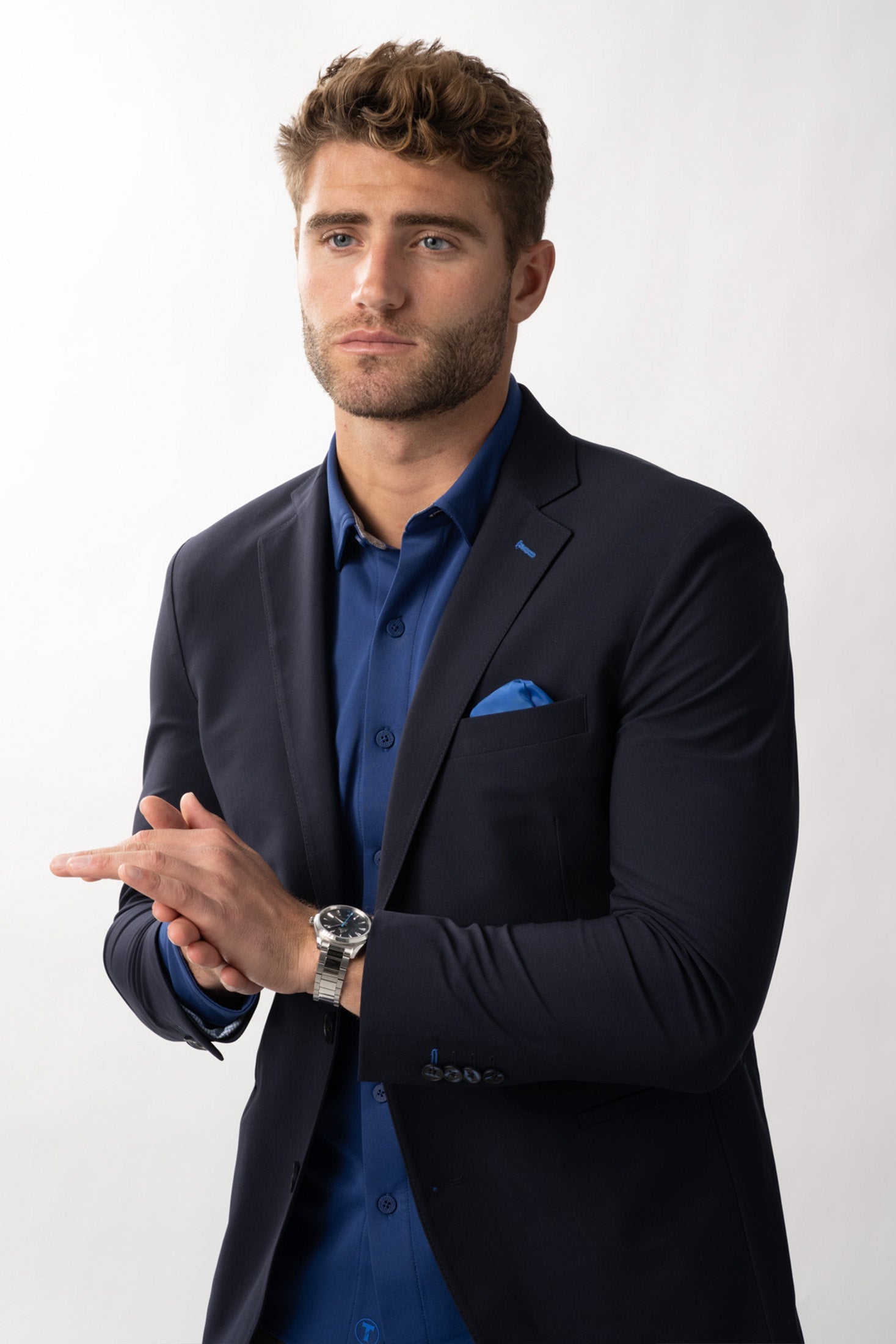 A young man with short brown hair and a trimmed beard wears the Twillory Performance Blazer // NAVY BLUE CONTRAST over a blue dress shirt with a blue pocket square and silver watch, standing against a white background, hands clasped.