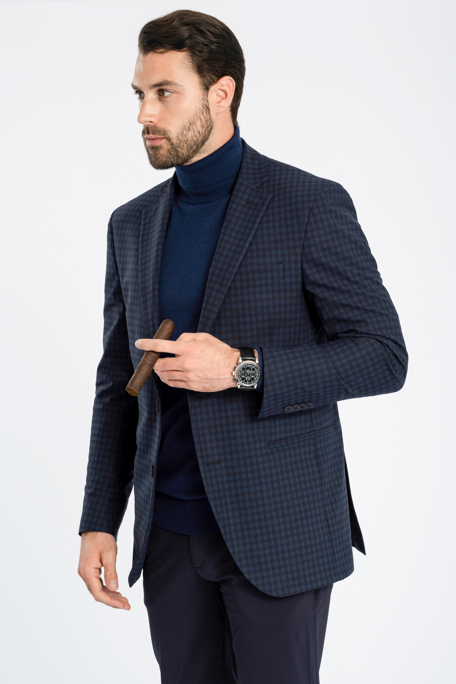 A man with dark hair and a beard wears Twillory’s Performance Blazer in Navy Check over a navy turtleneck and dark pants. He holds a cigar, sports a watch, and stands against a white background, gazing to the side.