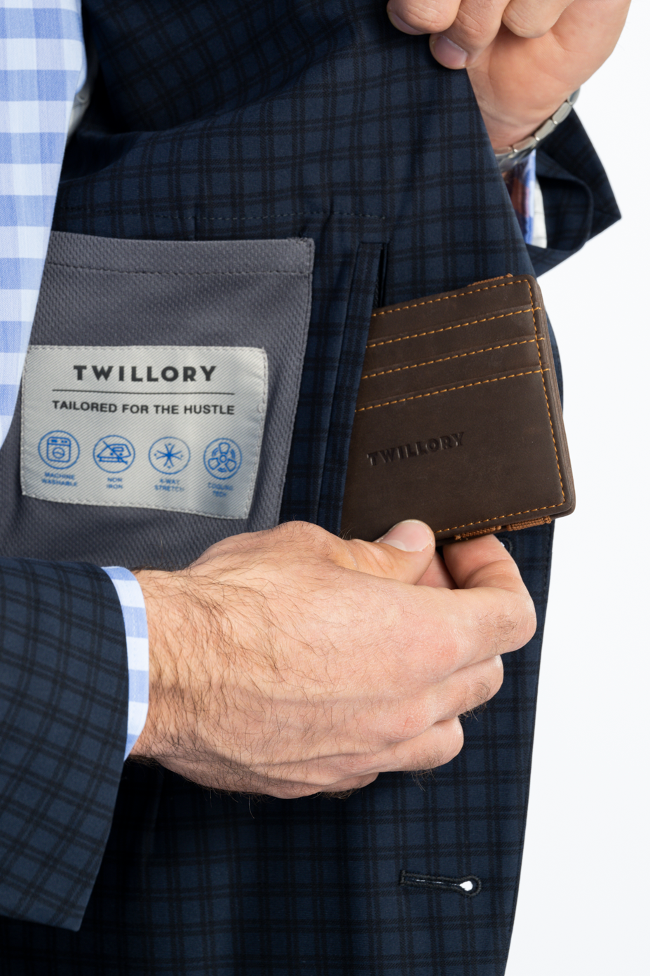 A man in a Twillory Performance Blazer // Navy Check and blue checkered shirt holds a brown leather card holder labeled “Twillory” inside the blazer’s pocket; the lining tag says “Twillory Tailored for the Hustle.”.