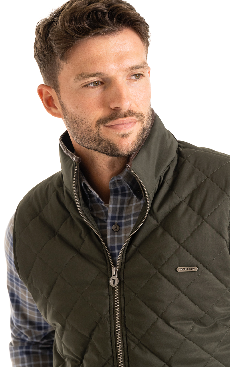 Man wearing a quilted green vest over a plaid shirt, looking to the side with a thoughtful expression.