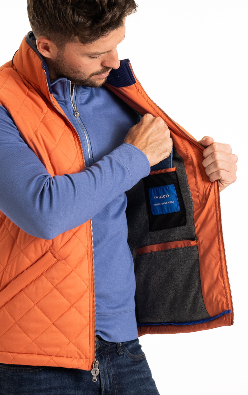 Man wearing an orange quilted vest opening the jacket to reveal the interior pocket with a blue label.