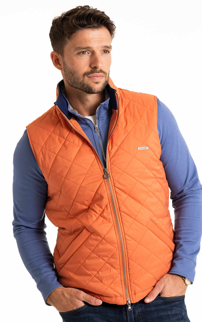 Man wearing orange quilted vest over blue long-sleeve shirt with hands in pockets.