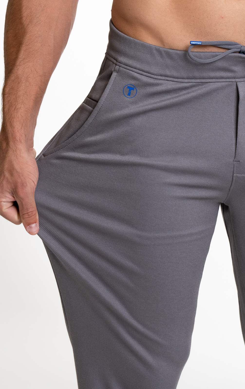 A person wearing Twillory’s Athlounger Jogger in gray stretches the fabric at the hip to show its flexibility, pulling it outward with their left hand. Their bare torso and right arm are visible, with a small blue logo near the pocket.