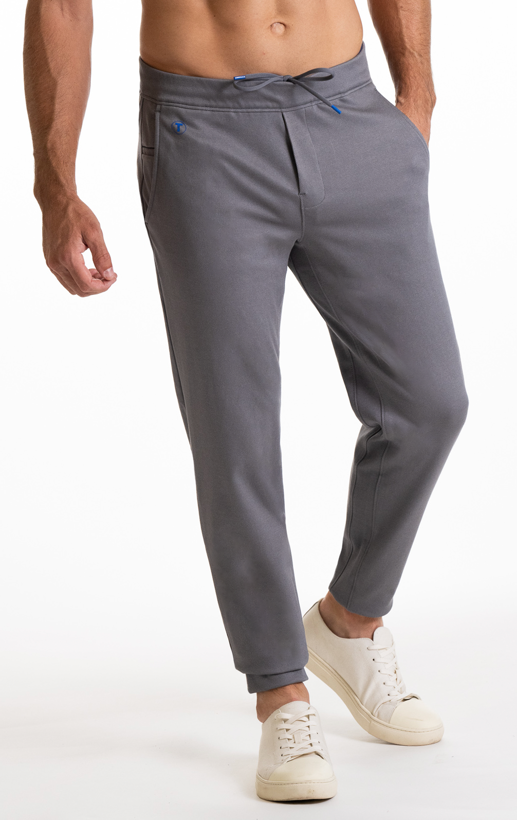 A man models the Twillory Athlounger Jogger—gray, tapered fit with an adjustable drawstring, blue accents, and side-zip pocket. He wears white sneakers, hands in pockets, torso visible against a plain white background.